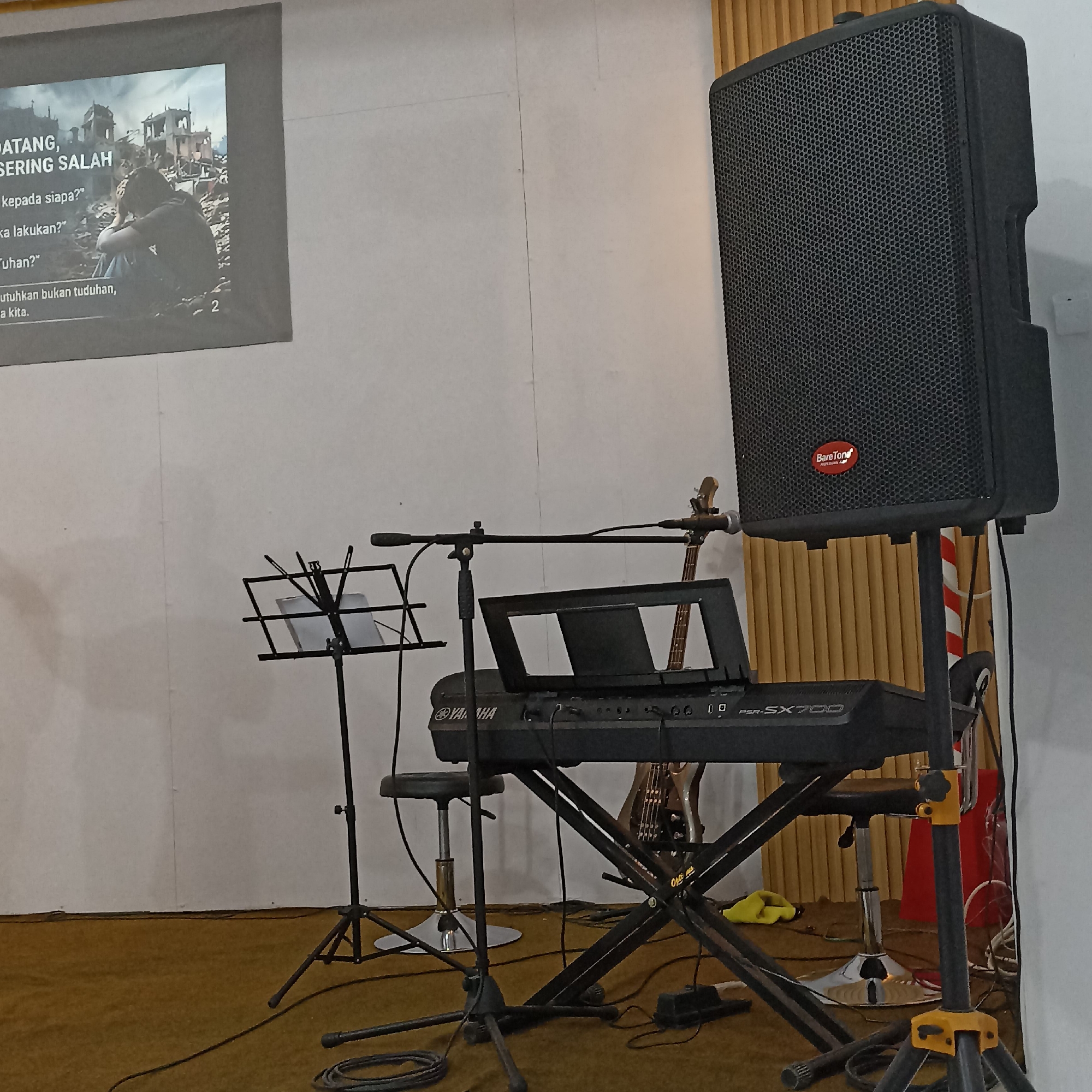 The image depicts an indoor setup, likely a stage or rehearsal space, featuring musical instruments and audio equipment. A large black speaker stands prominently on the right side, mounted on a tripod stand. In the center, a Yamaha PSR-SX700 keyboard is placed on a black X-style stand, with a music stand in front of it holding what appears to be blank paper. A microphone is mounted on a boom stand positioned over the keyboard. Behind the keyboard, the neck and body of an electric guitar are visible. A chrome-legged stool is situated to the left of the keyboard. In the background, a projection screen displays an image of a person sitting amidst ruined buildings and text in what appears to be Indonesian. The floor is covered in a brown carpet, and a white wall serves as the backdrop, with vertical wooden blinds visible on the far right. The scene suggests a setting for a musical performance or practice, possibly in a church or community hall. There are no people visible in the image. The overall impression is one of readiness for a musical event.