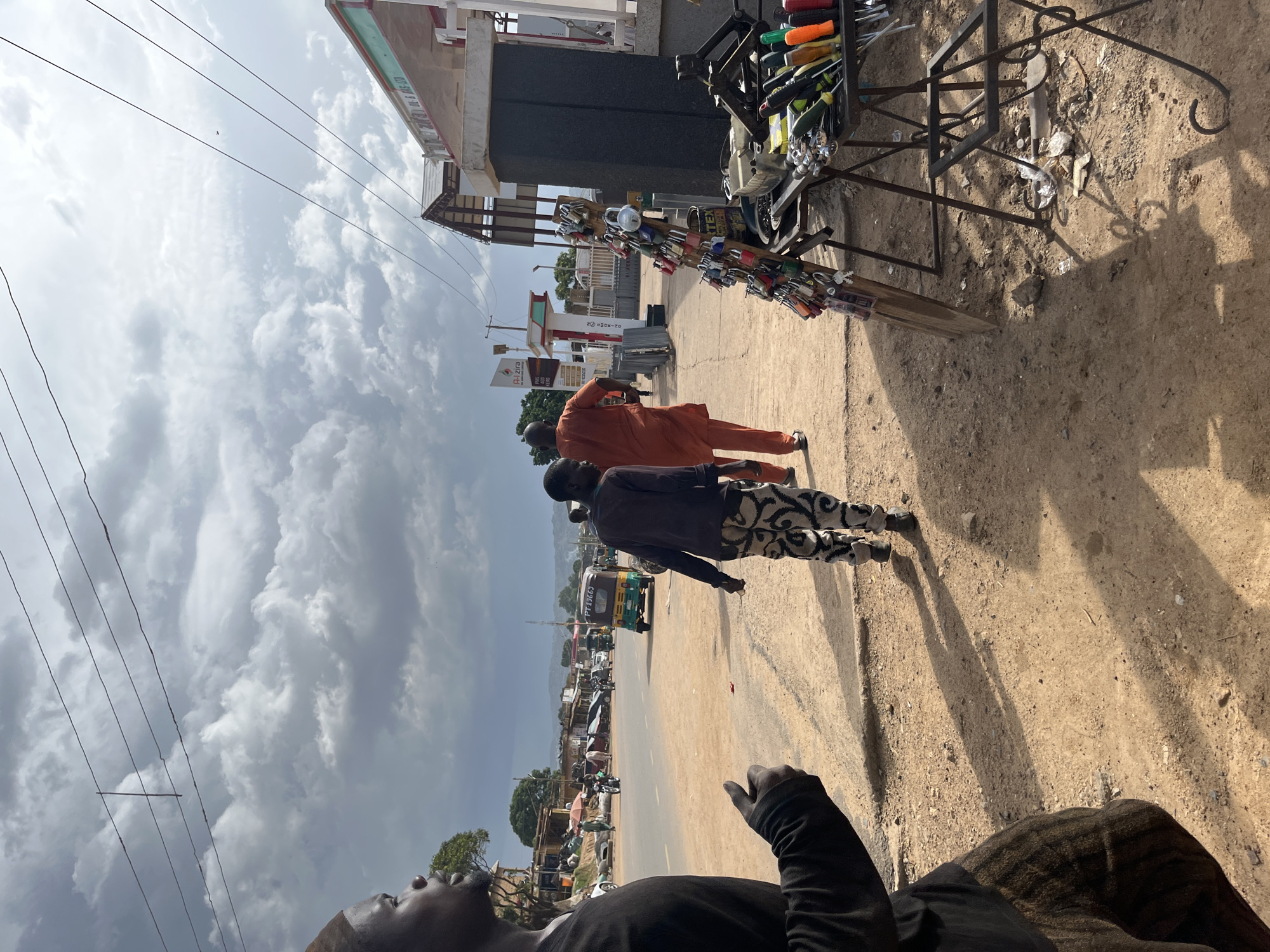 In Jos, Nigeria, a sunlit street scene unfolds. A man wearing a black long-sleeved shirt and a striped skirt walks away from the camera, his shadow stretching long across the dirt ground. To his right, another man in an orange outfit also walks away, his face obscured. In the background, a busy road is visible with several vehicles, including a yellow and green auto-rickshaw. Shops and buildings line the street, and a stall displays a variety of padlocks and tools. Overhead, power lines stretch across a cloudy sky. The overall impression is of a lively urban environment with a mix of commerce and daily life.