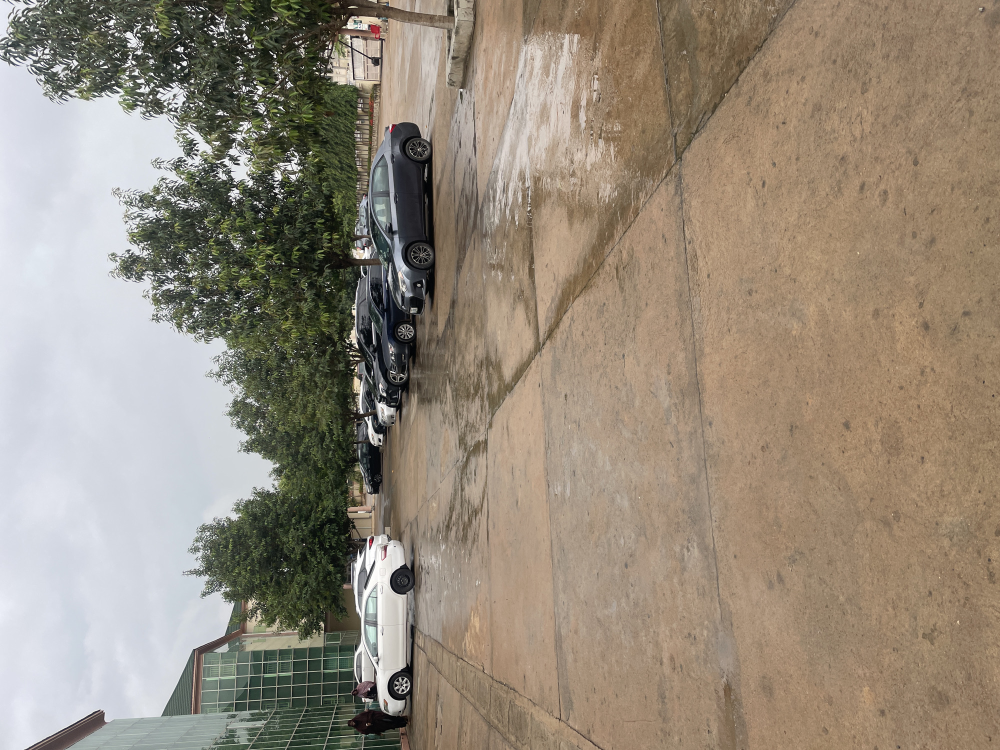 The image depicts a parking lot in front of a modern building with a glass facade, situated in Abuja, Nigeria. The ground is wet and reflects the overcast sky and surrounding trees, indicating recent rainfall. Several cars are parked in rows, including a white sedan on the left and various dark-colored sedans and SUVs further back. Two individuals, dressed in dark, modest clothing, stand near the white car on the left side of the frame. Trees line the edge of the parking lot, separating it from what appears to be a landscaped area and another building in the background. The overall scene is one of a quiet, possibly post-rainy day in an urban environment.