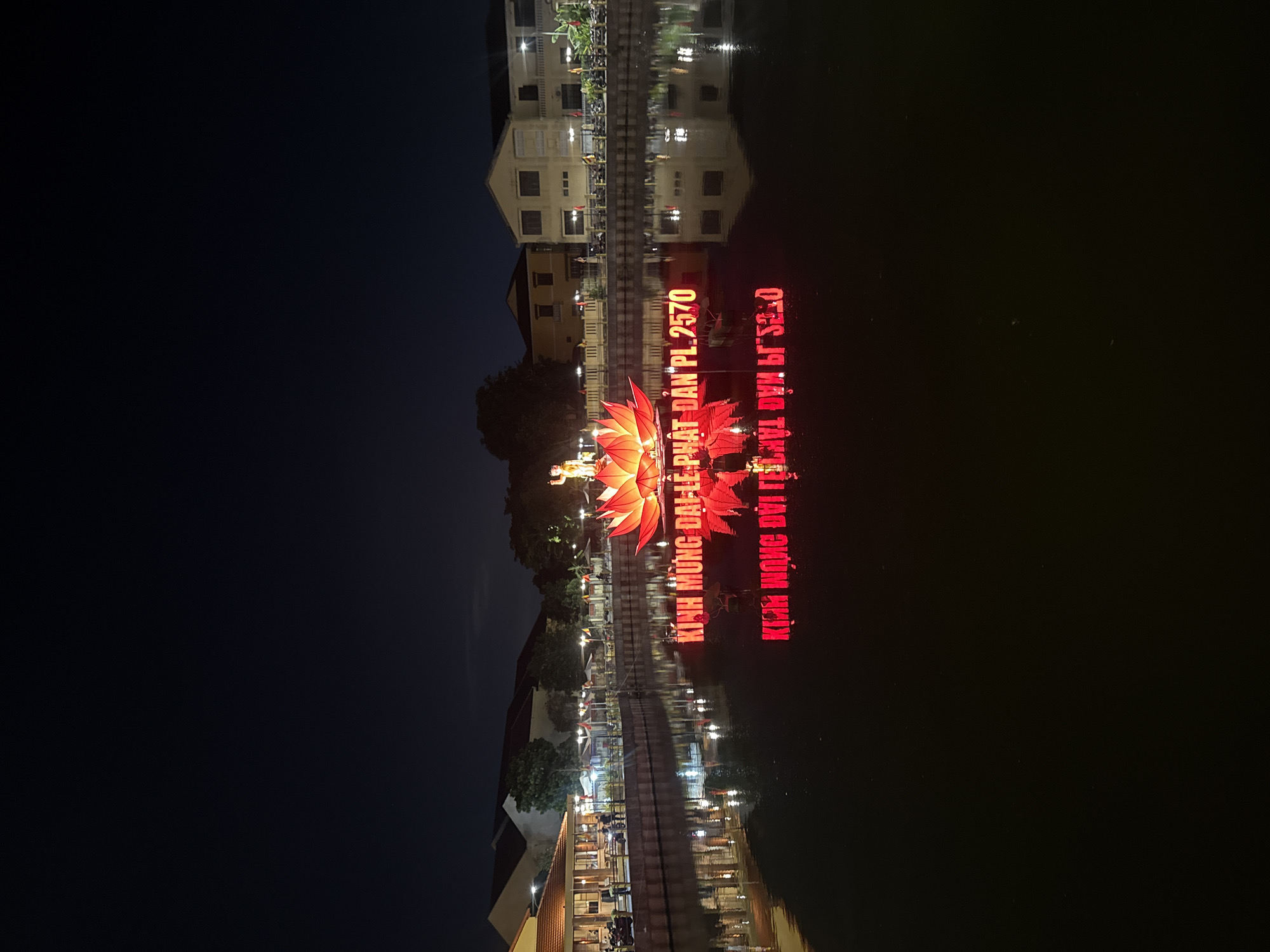At night, a body of water reflects buildings and illuminated displays, somewhere in Unknown, Unknown. The reflection showcases architectural elements like windows and rooftops. In the middle of the water, vibrant red lettering and a floral decoration stand out, also mirrored in the water. To the left of the water's edge, the night sky appears.