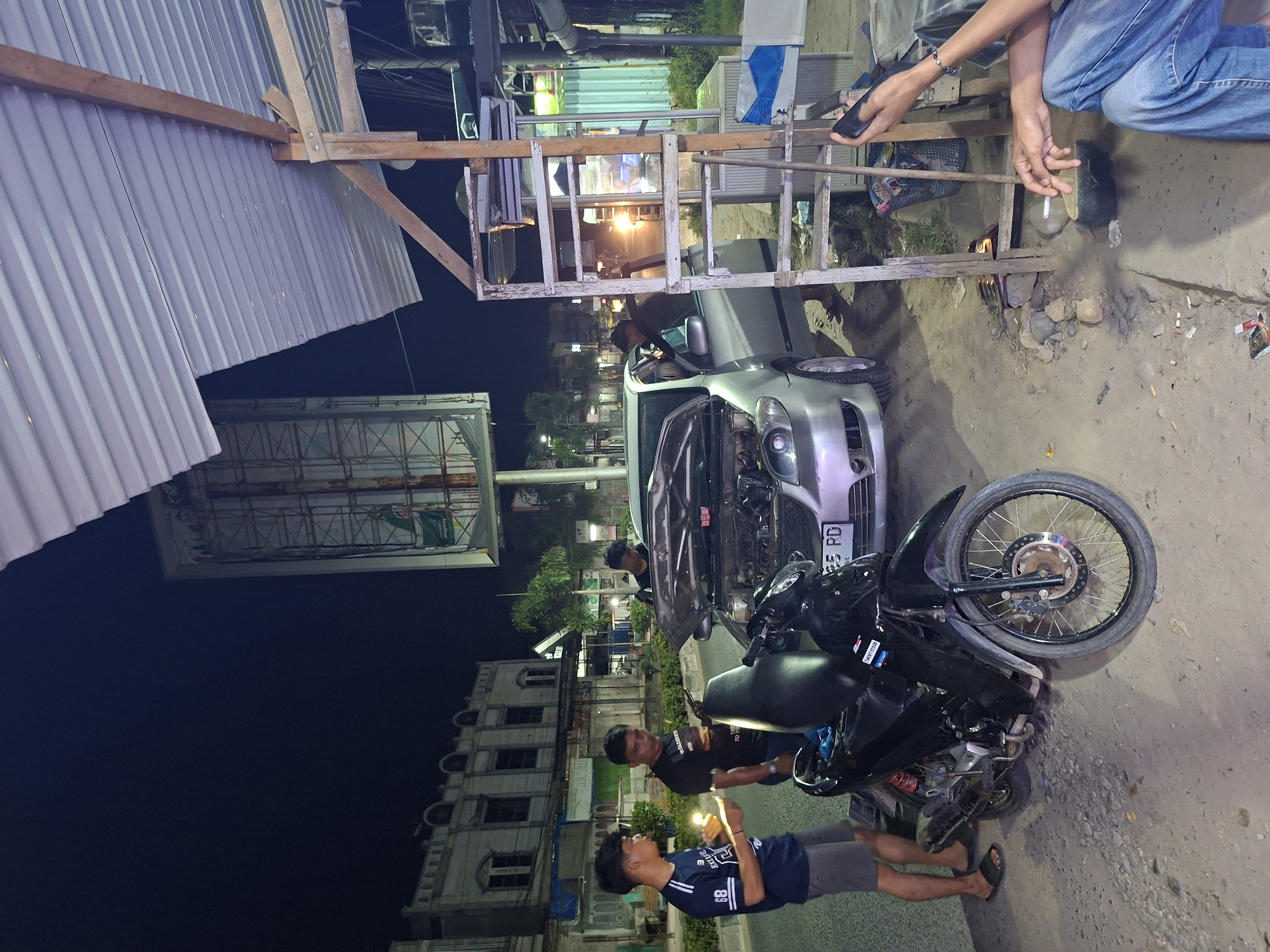 The image depicts an outdoor scene in Lhokseumawe, Indonesia, at night. A silver car with its hood open is parked next to a black motorcycle lying on its side. Three young men are standing near the motorcycle, seemingly examining the situation. The background includes buildings with a multi-story white facade, street lights, and trees. A makeshift wooden structure supports a corrugated metal roof on the left side. Overhead, a wooden frame supports some construction materials. The ground is a mix of dirt and gravel. A person's arm and hand are visible at the top edge of the image, holding a cell phone.