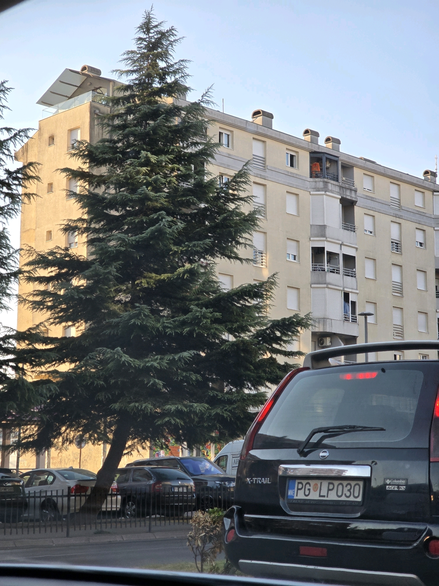 The image depicts an urban scene in Podgorica, Montenegro, with a focus on a street view dominated by a large evergreen tree and an apartment building. In the foreground, the rear of a black Nissan X-Trail SUV is visible, its license plate reading "PG LP 030". The SUV is parked behind a black metal fence. Other vehicles, including a silver sedan and a dark-colored wagon, are parked further down the street, also behind the fence. The apartment building, painted in a light yellow hue, features multiple floors with balconies and windows, many of which are covered by white rolling shutters. One balcony on a higher floor has an orange garment draped over a chair. The sky is clear and blue, suggesting daytime. The overall impression is of a residential area in Podgorica with typical urban elements.