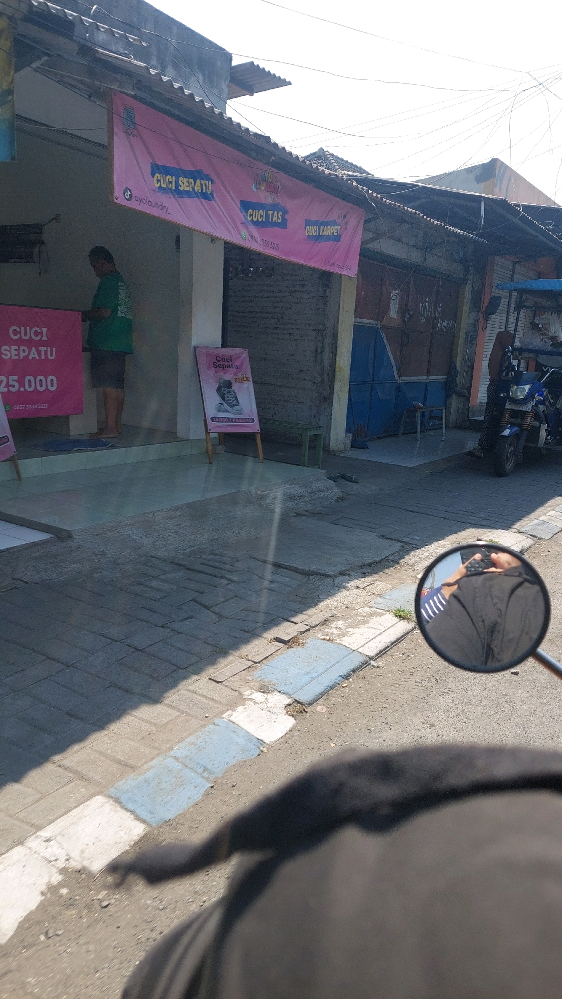 In the city of Sidoarjo, Indonesia, a street view is captured, featuring a storefront with prominent pink banners advertising laundry services. One banner, hung across the building, displays "CUCI SEPATU" (shoe cleaning), "CUCI TAS" (bag cleaning), and "CUCI KARPET" (carpet cleaning). Below the "CUCI SEPATU" text on this banner, a TikTok handle "@ayola_laundry_" is visible, along with a phone number that appears to be "0896 333 3213". Another banner on the left side of the storefront, also pink, reiterates "CUCI SEPATU" and displays a price of "25.000" with a phone number below it. A standing sign on the right of the entrance shows an image of a shoe and the text "Cuci Sepatu" and "25.000 / PASANG".
A man in a green t-shirt and dark shorts is standing inside the storefront, appearing to be interacting with something on a counter. The entrance to the store has a tiled floor and a mat. A blue and white striped object, possibly a mat or a painted curb, runs along the edge of the sidewalk.
In the foreground, a motorcycle mirror reflects a person wearing a dark shirt, and partially visible is a person's arm. The road surface is asphalt, with some tiled areas and concrete slabs visible along the sidewalk. The background shows a building with a corrugated roof and shuttered windows, and a blue-doored structure. A blue and white motorized scooter is parked on the right side of the image. The scene is illuminated by sunlight, casting shadows on the ground.