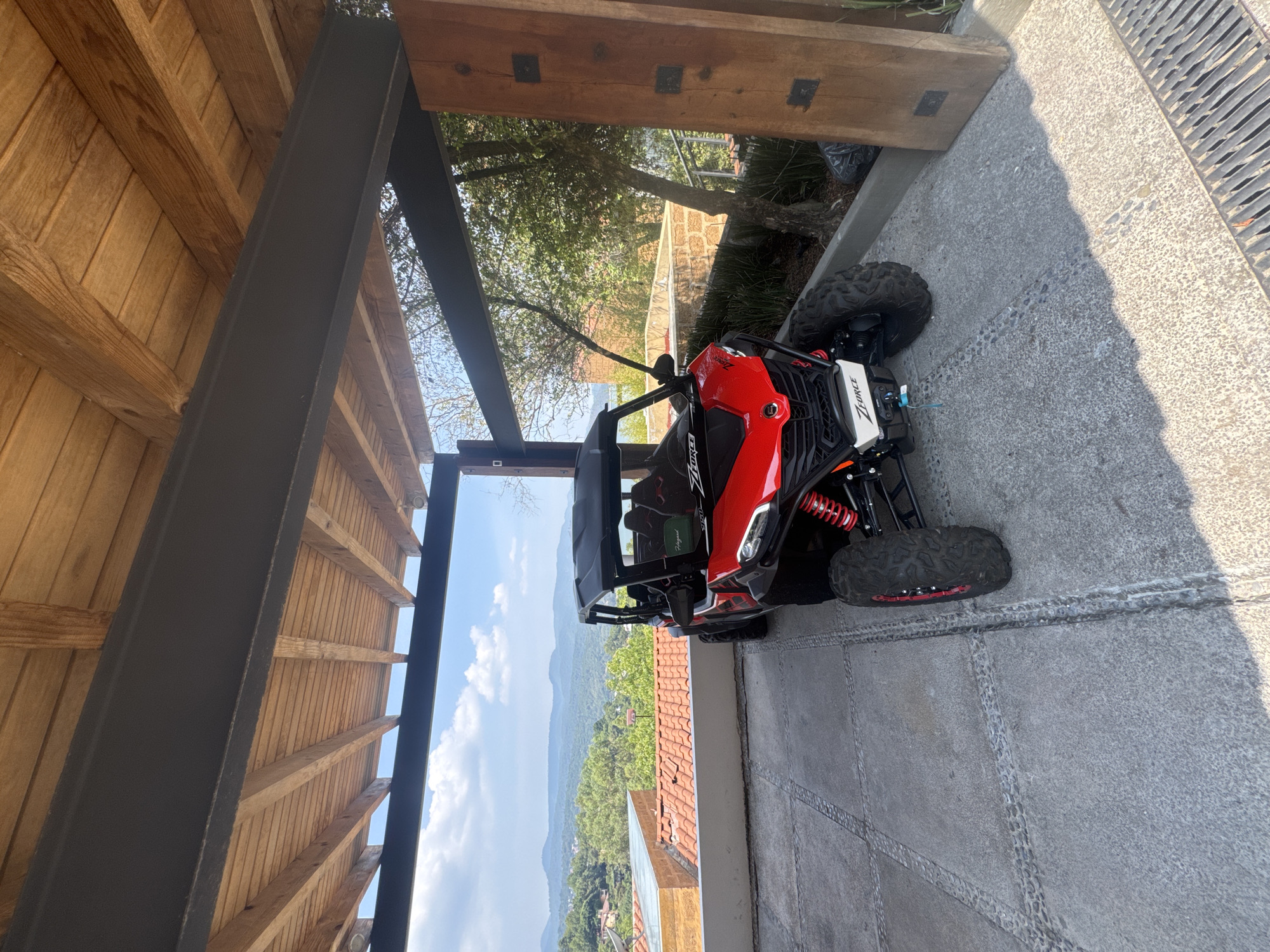 The image shows a red and white Z-Force side-by-side vehicle parked on a concrete surface with tire tracks, possibly a driveway or patio. The location is suggested to be Valle de Bravo, Mexico, based on the view of mountains and a landscape with red-tiled roofs visible in the background. The vehicle is positioned close to a building with an overhanging wooden roof supported by dark metal beams. A drainage grate runs along the edge of the concrete surface. There are no people visible in the frame.