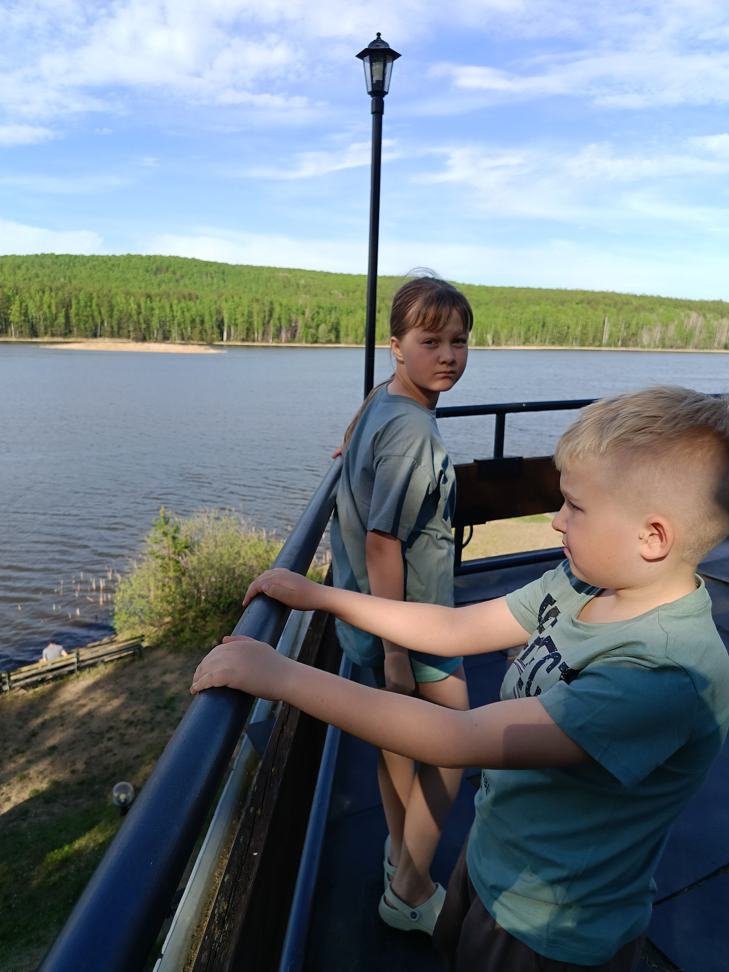 The image shows two children, a girl and a boy, standing on a balcony overlooking a lake and forest. The girl is standing behind the boy, looking towards the camera with a slightly serious expression. She has light brown hair pulled back in a braid, and she is wearing a greenish-blue t-shirt and shorts, and light colored shoes. The boy is in the foreground, facing to the right of the frame. He has blonde hair and is also wearing a similar colored t-shirt and brown shorts. He is holding onto the black metal railing of the balcony.
The balcony is made of wood and metal, with a black railing. A black lamp post stands behind the girl. Beyond the balcony, there is a large body of water, a lake, stretching to the horizon. On the opposite shore, there is a dense green forest. The sky is blue with some white clouds.
In the far background on the left side of the frame, there is what appears to be a small seating area or dock with people sitting there. Based on the context clues of the image, the location is likely in or near Chaykovsky, Russia, given the mention of the city. The environment suggests a recreational or scenic area near a lake.