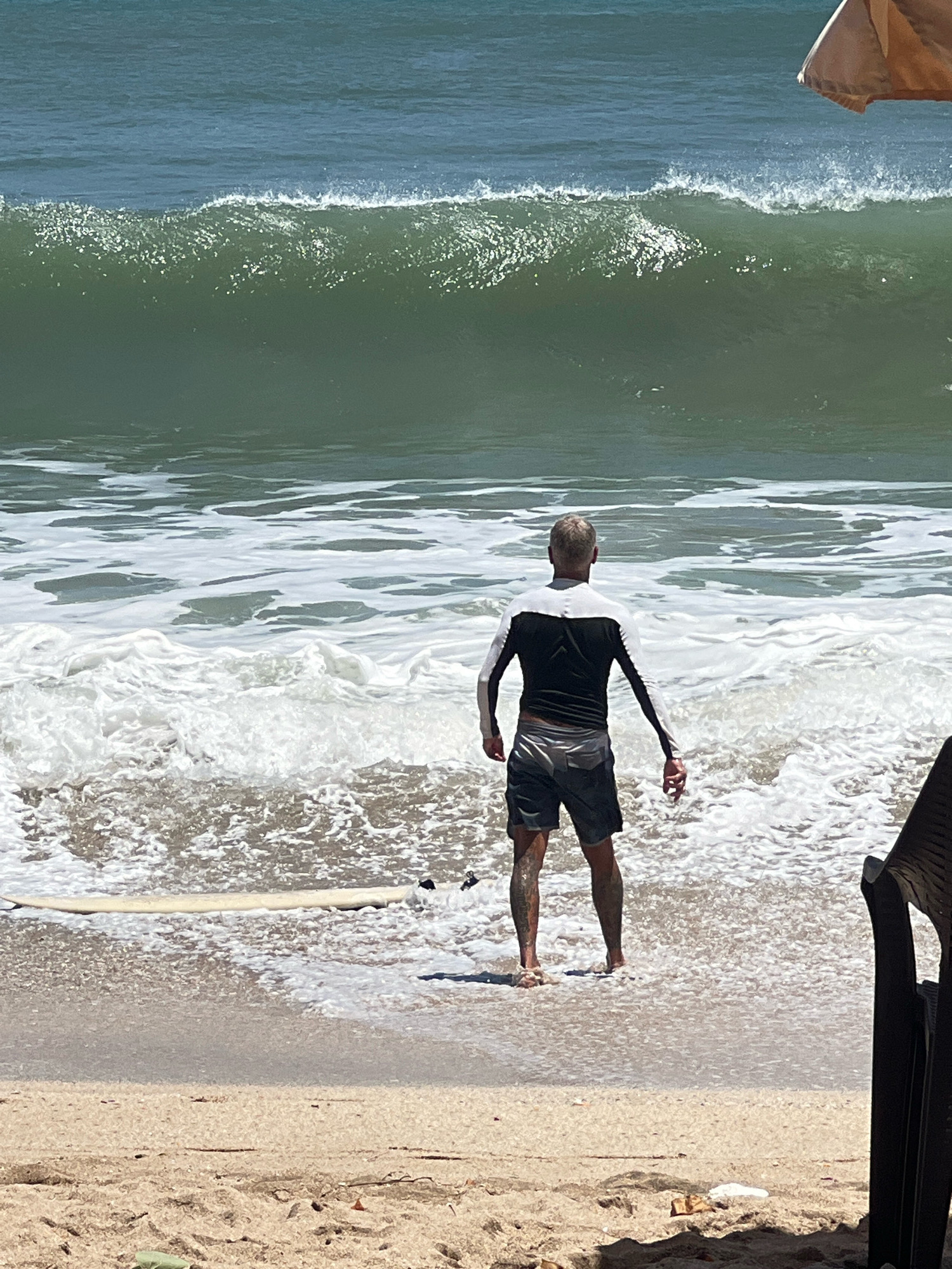 A man stands facing the ocean on a sandy beach in Kuta, Indonesia. He is wearing a long-sleeved rash guard with black and white panels, and dark shorts. The man's legs are wet, and he appears to be standing in shallow water or surf. His right hand is extended slightly, holding what appears to be a small, light-colored object. A surfboard is visible on the sand to his left, partially submerged in the water. In the background, a large, green wave is breaking, with white foam and churned water spreading towards the shore. The sky is clear and bright, suggesting a sunny day. The overall scene depicts a person about to enter or having just exited the ocean for surfing or swimming.