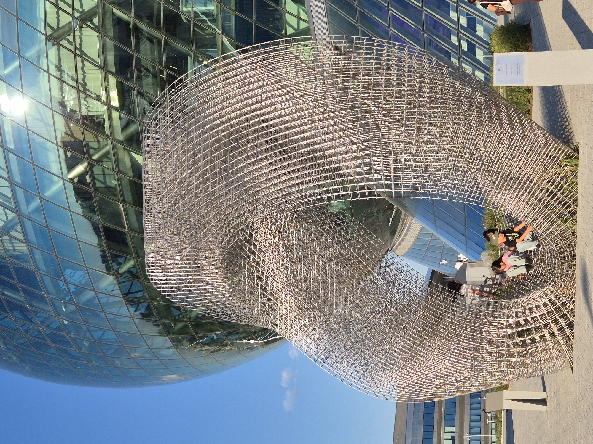 The image shows an outdoor scene in Astana, Kazakhstan, with a prominent spherical glass building in the background. In the foreground is a large, intricate sculpture made of metal mesh, forming a hollow, twisted structure. Three children are visible inside the sculpture, two girls and one boy, appearing to be sitting and interacting. The boy is wearing a green baseball cap and a black t-shirt with a graphic. One of the girls is wearing a pink shirt and blue jeans, and the other appears to be wearing a white top and dark bottoms. The sculpture is set against a paved plaza with some low-lying vegetation and a signpost to the right. The sky is clear blue with a few scattered clouds. The overall impression is of a public space featuring modern architecture and a striking art installation.