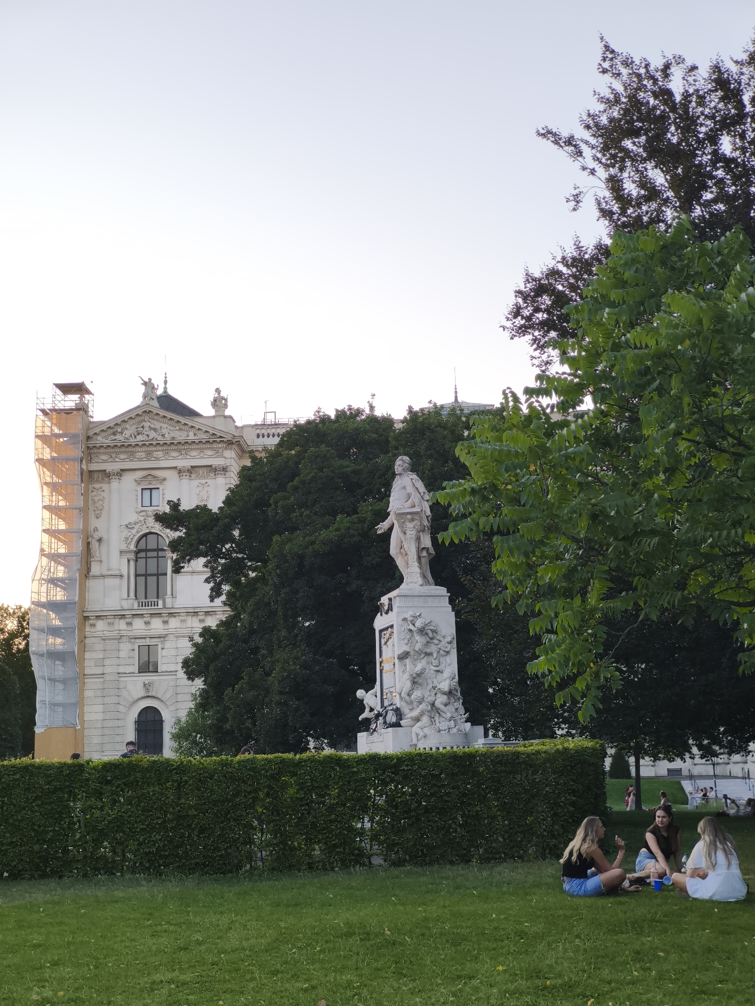 The image depicts the Monument to Wolfgang Amadeus Mozart, a prominent landmark located in the Burggarten park in Vienna, Austria. In the foreground, three young women are seated on the grass, engaged in conversation. Behind them, a meticulously maintained green hedge separates the park area from a grand, ornate building, which appears to be undergoing renovations as evidenced by the scaffolding on its left side. The Mozart monument, a large white marble sculpture, stands tall behind the hedge, featuring Mozart himself in a distinguished pose atop an elaborate pedestal adorned with allegorical figures. Lush green trees frame the scene, with one large tree in the right foreground partially obscuring the view. The sky above is a clear, pale blue, suggesting it is late afternoon or early evening. Several other people are visible in the background, scattered throughout the park.