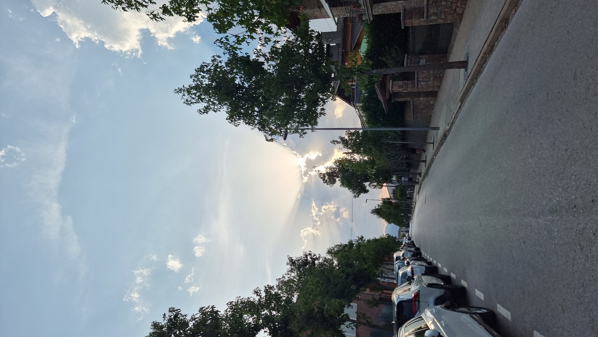 The image depicts a street scene in the city of Sant Joan Les Fonts, Spain, with a view down a residential road lined with parked cars and trees. The sun is setting, with its rays breaking through the clouds and casting a dramatic light across the sky. The road is asphalt and appears to be in good condition, with white dashed lines marking the parking spaces on the left side. Several white cars are parked along the curb. On the right side of the road, there is a sidewalk and a stone wall with an arched entrance. A street lamp is visible, as are houses and buildings in the background. Two individuals are walking on the sidewalk. The overall impression is a tranquil evening in a quiet neighborhood.