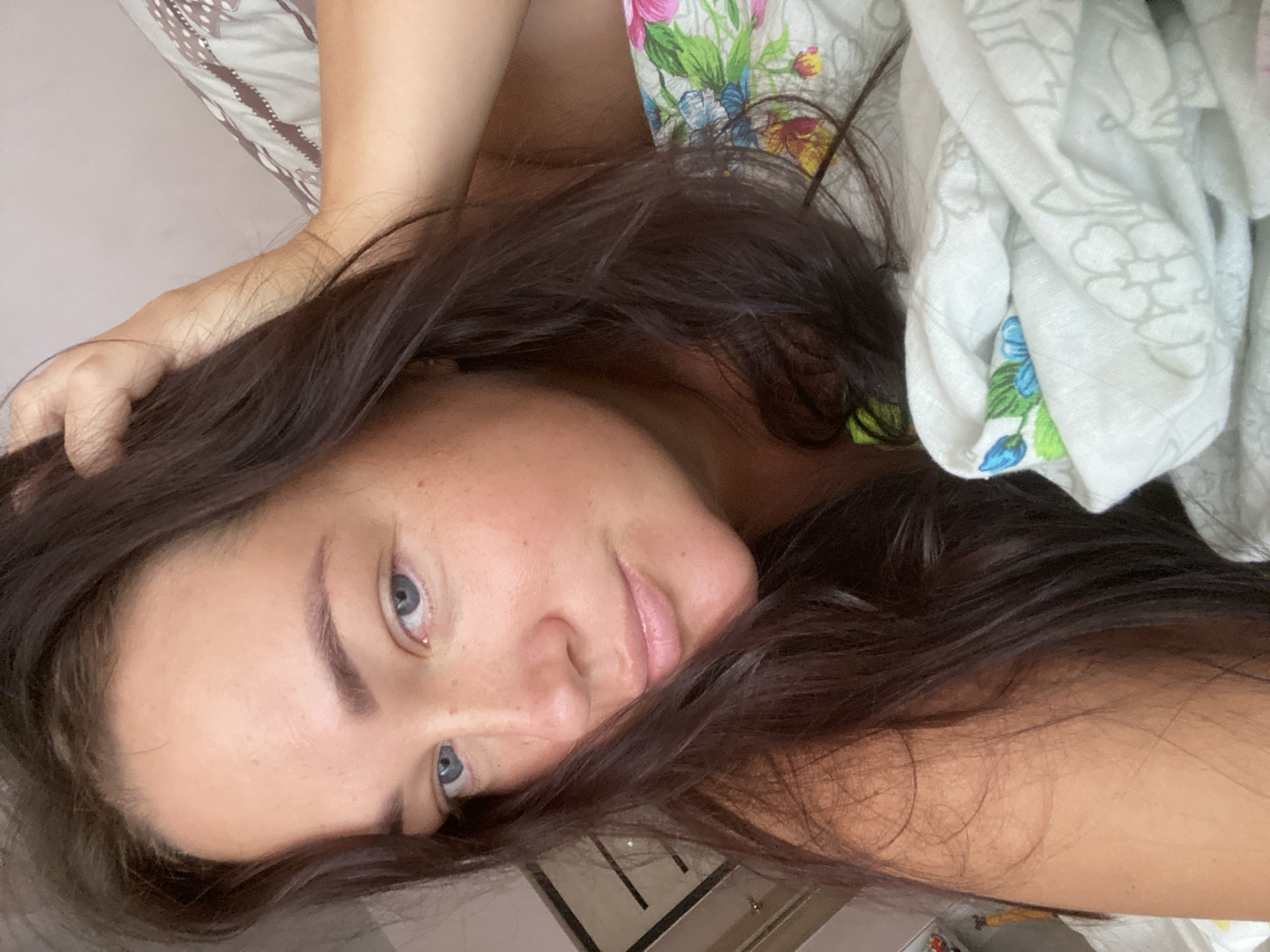 The image is a close-up, selfie-style shot of a woman with long, dark brown hair and blue eyes, lying in bed. She is wearing a green top and is partially covered by a floral-patterned duvet. Her right hand is resting on her head, and her left arm is also visible. The background shows a pinkish wall and a dresser with a framed picture. The lighting appears to be natural, possibly from a window. The overall impression is that of a casual, intimate moment. The image is not geotagged and there is no information to suggest the location is Sochi, Russia.