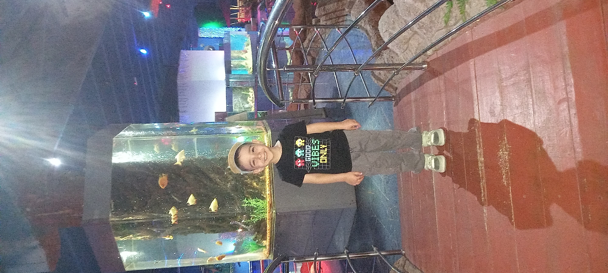 A young boy stands next to a tall aquarium filled with fish, indoors. He wears a dark shirt with "GOOD VIBES ONLY" printed on it, grey pants, and light-colored shoes. He also wears a light-colored hat and is smiling. The aquarium contains various small, golden fish. Other aquariums are visible in the background, along with metal railings and a section of stone-like wall. The flooring is red and wooden. The lighting is mixed with blues and reds in the background. The location is likely in Krasnodar, Russia based on the context.