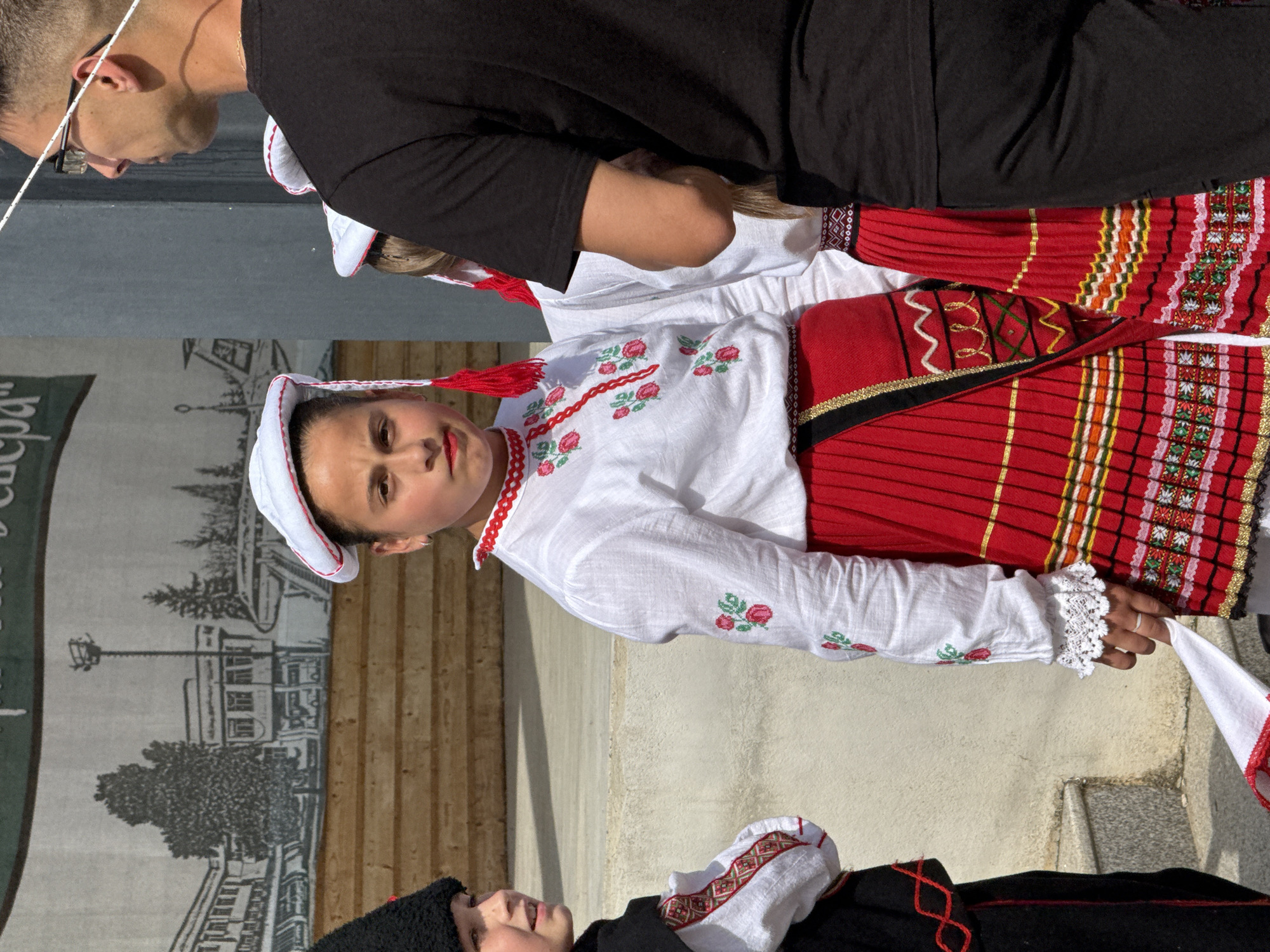 The photograph captures a cultural event in the city of Pordim, Bulgaria, likely a folk festival or performance. In the foreground, a young girl dressed in traditional Bulgarian attire stands prominently. Her costume consists of a white embroidered blouse with red floral motifs, a red pleated skirt with intricate woven patterns, and a white headdress adorned with red tassels. To her left, another child in a black and red traditional outfit is partially visible. Behind them, a man wearing glasses and a black t-shirt observes the scene. The backdrop features a wooden stage with a painted mural depicting buildings and trees. The overall atmosphere suggests a community gathering celebrating local heritage.