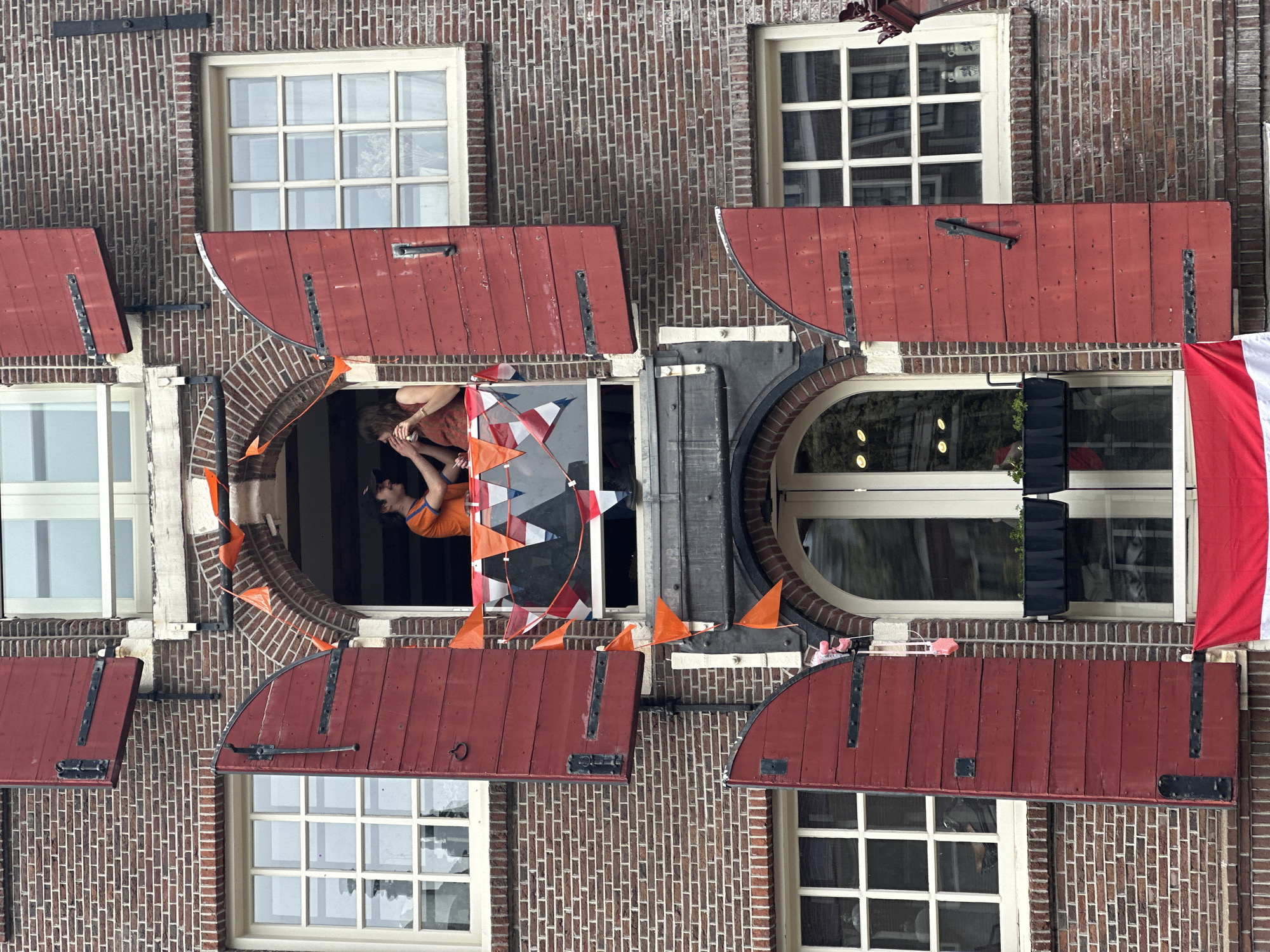 The image displays a festive scene on the brick facade of a building, typical of Amsterdam architecture. Several windows with white frames and traditional red wooden shutters are visible across the brown brickwork; some shutters are open, others closed.
In the central part of the image, two individuals are leaning out of an open window. One person, wearing an orange top and a dark cap, is clearly visible, looking outwards. A second person is partially obscured beside them. The window they are leaning from is adorned with strings of triangular bunting featuring orange, white, and blue flags. Additional small, individual orange flags are also placed around the window frame and on the brickwork above adjacent shutters.
Below a closed red shutter on the right side of the frame, a Dutch flag, characterized by its vertical red, white, and blue stripes, hangs prominently. The lighting suggests it is daytime, with soft or overcast conditions and no harsh shadows. The widespread use of orange decorations and the presence of the Dutch national flag strongly indicate a celebration or national event, such as King's Day (Koningsdag) or a major sporting occasion, taking place in the Netherlands. There is no visible text in the image.