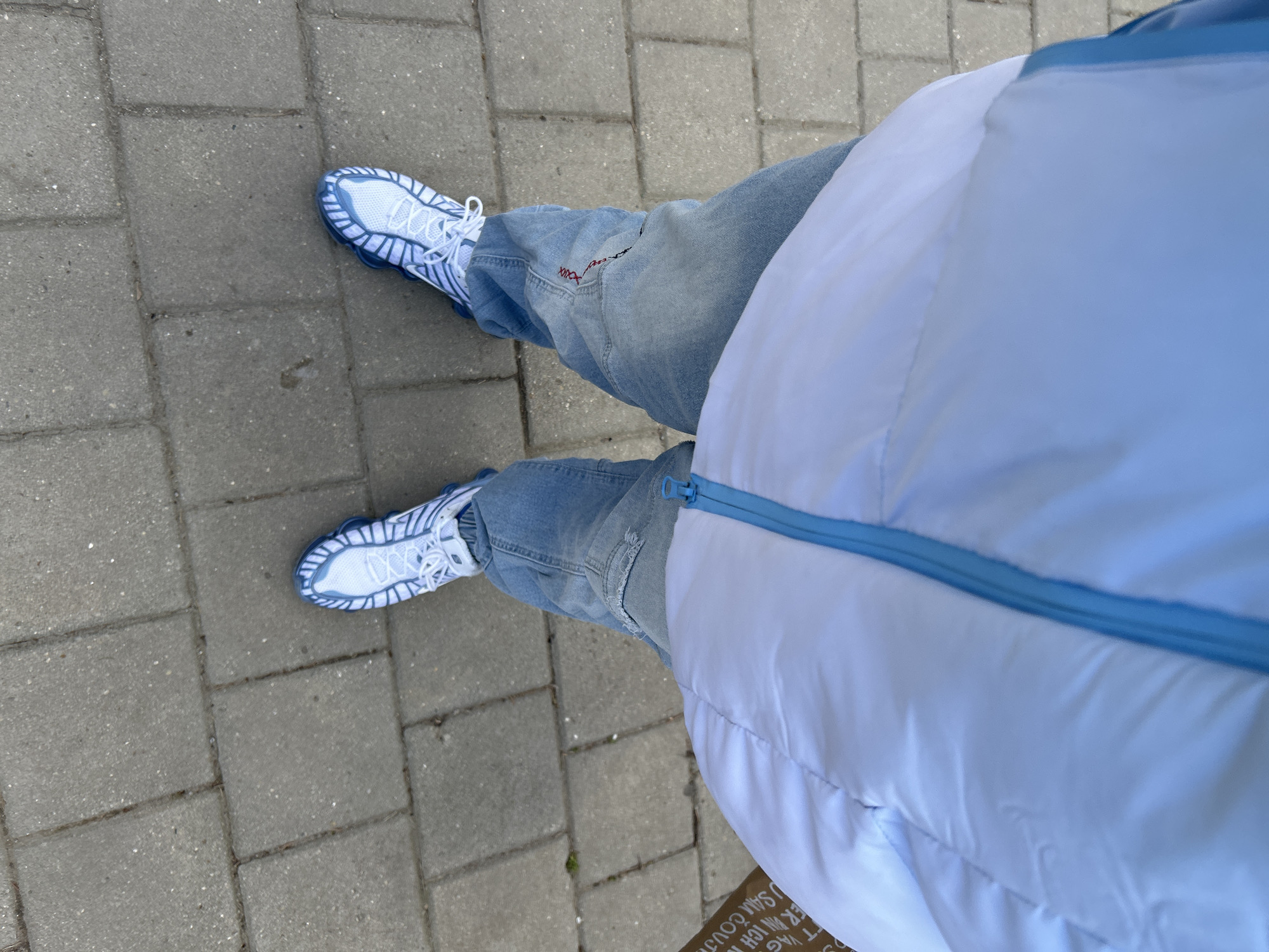 The image shows a person standing on a sidewalk made of gray rectangular paving stones. The person is wearing light blue jeans and white and light blue sneakers with a distinctive patterned sole. They are also wearing a puffy white vest with a light blue zipper. The view is from above, looking down at the person's feet and clothing. At the bottom right corner of the image, a small portion of a box with text in German is visible. The location appears to be an urban setting, likely a city street, but without additional context, it is difficult to confirm whether the location is Sofia, Bulgaria.