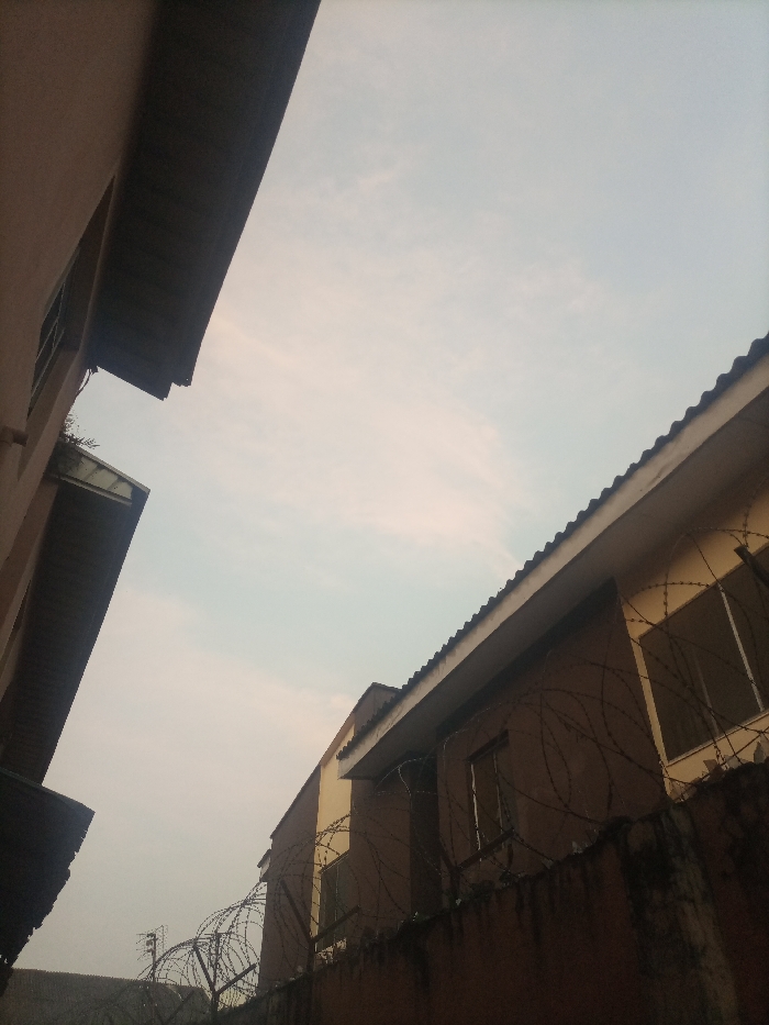 A low-angle shot features two buildings against a muted sky. The building on the left displays a dark overhang. The building on the right is brown with windows visible and barbed wire along the top of the wall. The overall scene seems hazy.