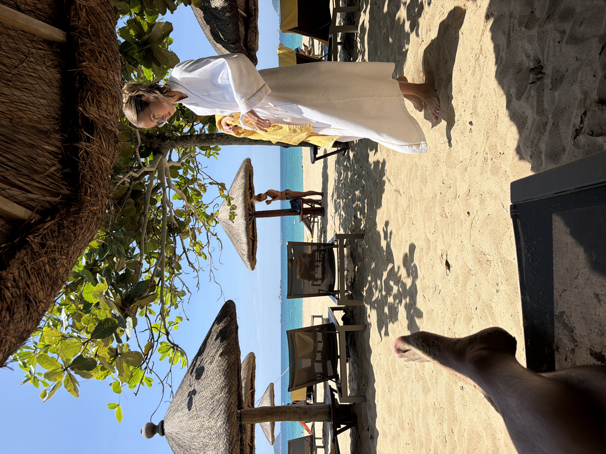 The image depicts a beach scene under a thatched umbrella. In the foreground, a person's bare foot is visible, resting on the sand. To the right, a woman in a white bathrobe is holding a yellow towel and looking downwards. Behind her, the ocean is visible with a clear blue sky. In the background, other beach umbrellas, lounge chairs, and people can be seen. The sand is light brown and there are shadows cast by the umbrella and trees. The overall setting suggests a tropical or subtropical beach resort. The location is identified as Jiyang District, China.