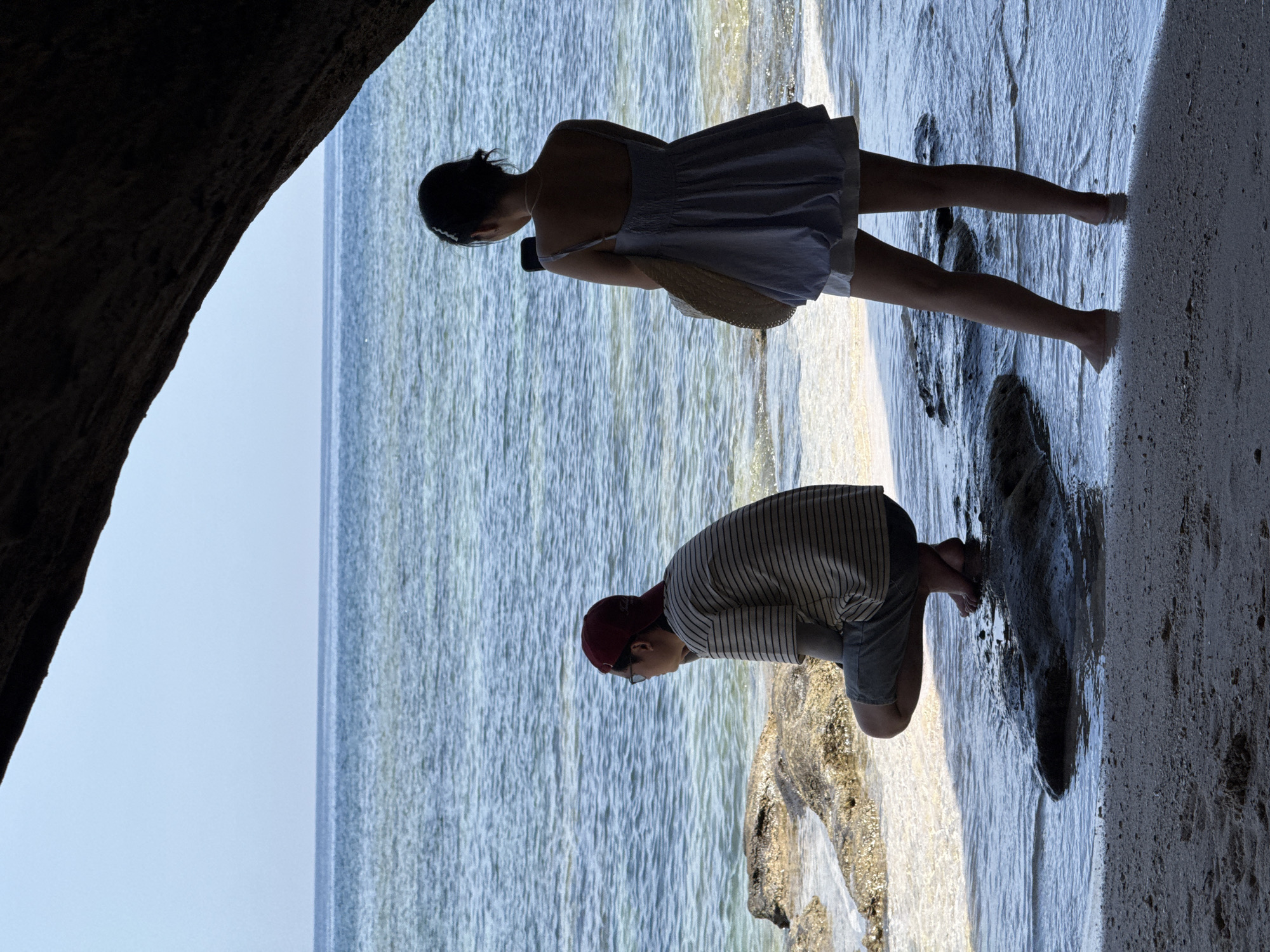 Two individuals, silhouetted against the bright sea and sky, stand on a rocky shore. The person on the left, wearing a striped t-shirt, shorts, glasses, and a red cap, is crouched down, looking out at the water. The person on the right, a woman in a light-colored dress and shorts, stands facing away from the camera, holding a smartphone to take a picture. She carries a straw bag over her shoulder. The water is clear and shallow, with sunlight reflecting off its surface and illuminating the rocks beneath. The foreground is a sandy beach, with a large, dark rock overhang creating a shadow over the scene. The location is a beach on Lý Sơn Island, Vietnam, a Special Administrative Region known for its volcanic landscapes and clear waters.