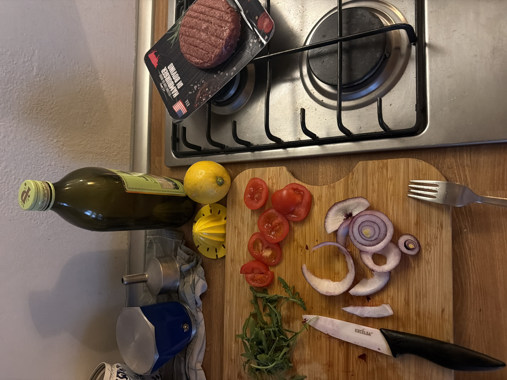 The image presents a top-down view of a kitchen scene, likely in Milan, Italy given the Italian text on the hamburger packaging. A stainless steel stovetop with two burners occupies the upper portion of the frame. A package of "Hamburger di Bovino" with an American flag is positioned precariously on the burner grate.
Below the stovetop, a wooden cutting board is the central focus. It holds sliced red tomatoes, several rings of red onion, and a small bunch of arugula. A yellow lemon juicer and a bottle of olive oil are to the left of the cutting board. A silver fork and a black-handled knife lay on the cutting board as well.
A blue moka pot, a silver funnel, and a dishtowel are seen hanging on the wall to the left of the olive oil bottle. The setting appears to be a home kitchen, characterized by ordinary, everyday objects used in food preparation. There are no people visible in the image.