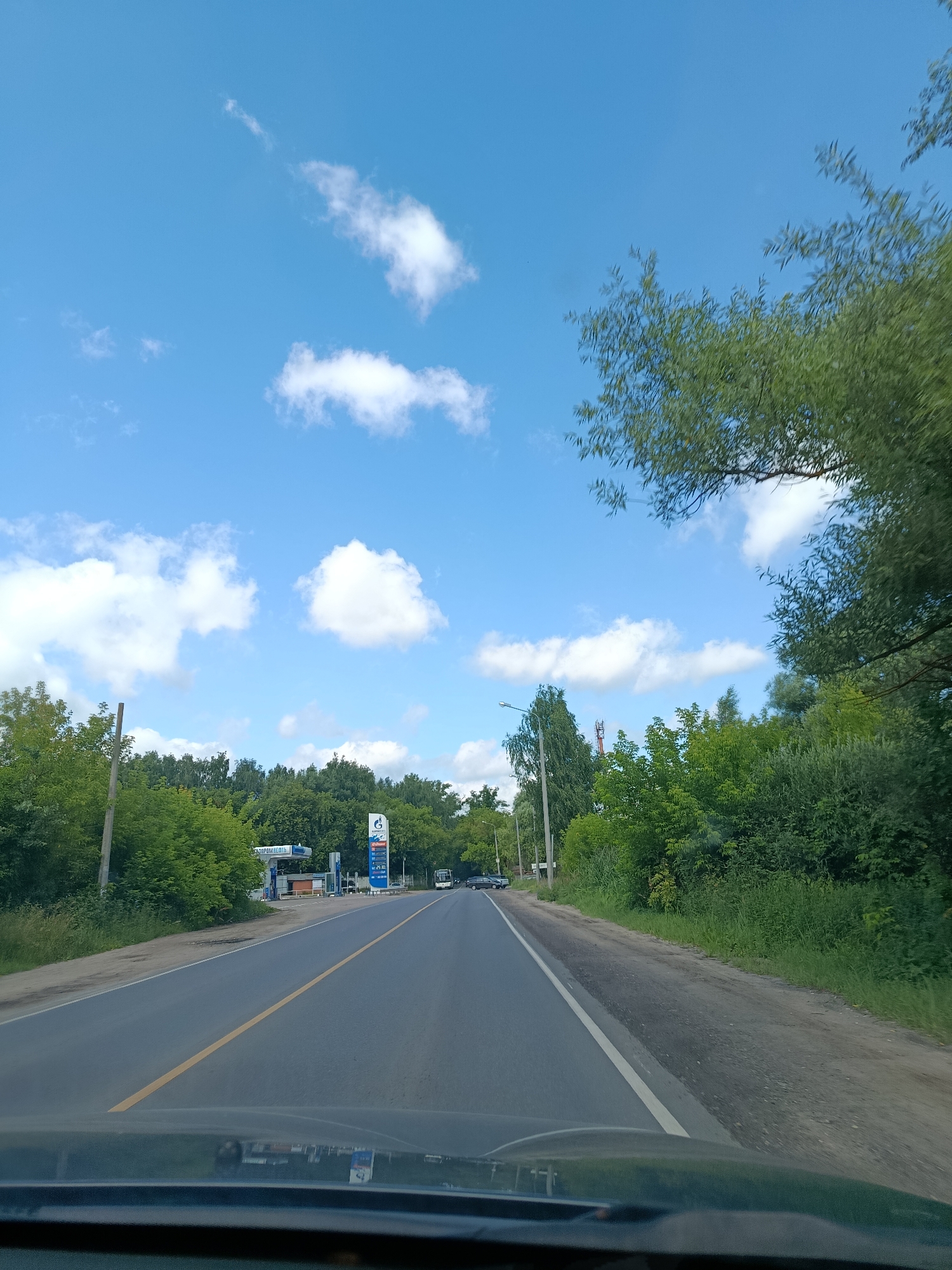 The image depicts a view from inside a vehicle traveling on a road in Orekhovo-Zuyevo, Russia. The road is paved with a yellow line down the center and a white line on the right edge. On the left side of the road, there is a gas station with a blue and white sign that reads "ГАЗПРОМНЕФТЬ" (Gazpromneft). Several cars are visible at the gas station, and a white bus is seen further down the road. The surrounding environment is characterized by lush green trees and foliage on both sides of the road. The sky is bright blue with scattered white clouds. The image appears to have been taken on a sunny day during the daytime.