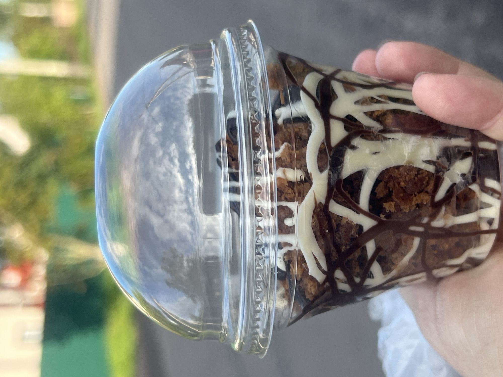 A close-up, eye-level shot shows a hand holding a clear plastic cup filled with a dessert. The dessert appears to be a layered confection of crumbled cake or cookie pieces, drizzled generously with both dark chocolate and white chocolate syrup. The cup is topped with a clear, domed plastic lid. The background is blurred, suggesting an outdoor setting in Otradnoye, Russia. The blurred elements include hints of green foliage, buildings, and a street. The overall impression is of a sweet treat being enjoyed outdoors, possibly on a street or in a park.