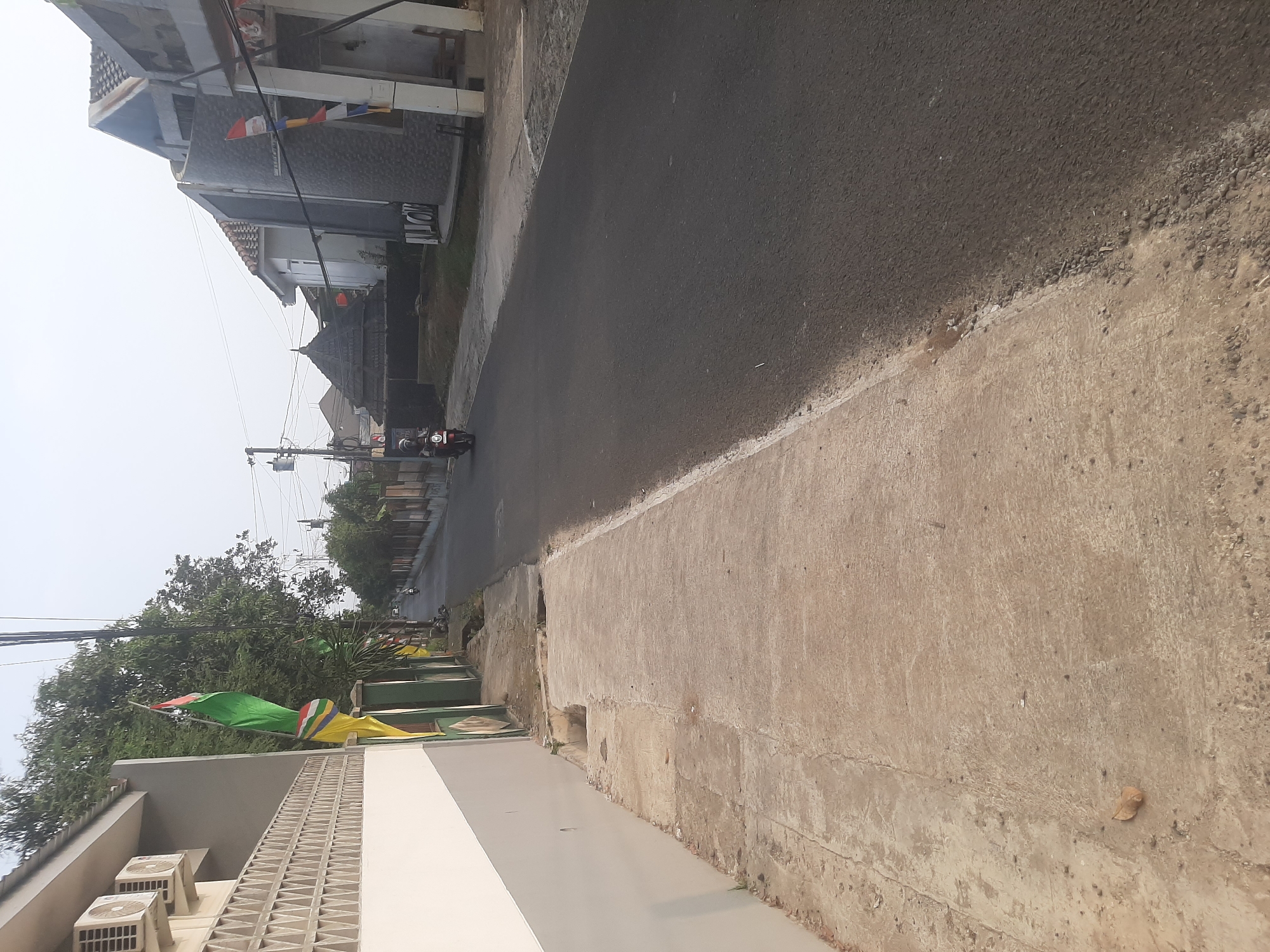 The image depicts a street scene in Sumampir, Indonesia. On the left side of the paved road is a sidewalk with a concrete surface. Adjacent to the sidewalk is a building with white and gray walls. An air conditioning unit is visible on the upper left wall. Several flags, including a green and red flag and a yellow flag with multicolor stripes, are attached to poles or structures along the sidewalk.
The road itself is dark asphalt, with a white line marking the edge. A single person is riding a red motorcycle in the distance, moving away from the viewer. On the right side of the road, there are several houses with tiled roofs and light-colored walls. Some of these houses have decorative flags or banners attached to their exteriors. Trees and vegetation are visible along the sides of the road and between buildings. The sky is overcast and appears hazy, suggesting it might be early morning or late afternoon. The overall scene is one of a residential street in a populated area.
