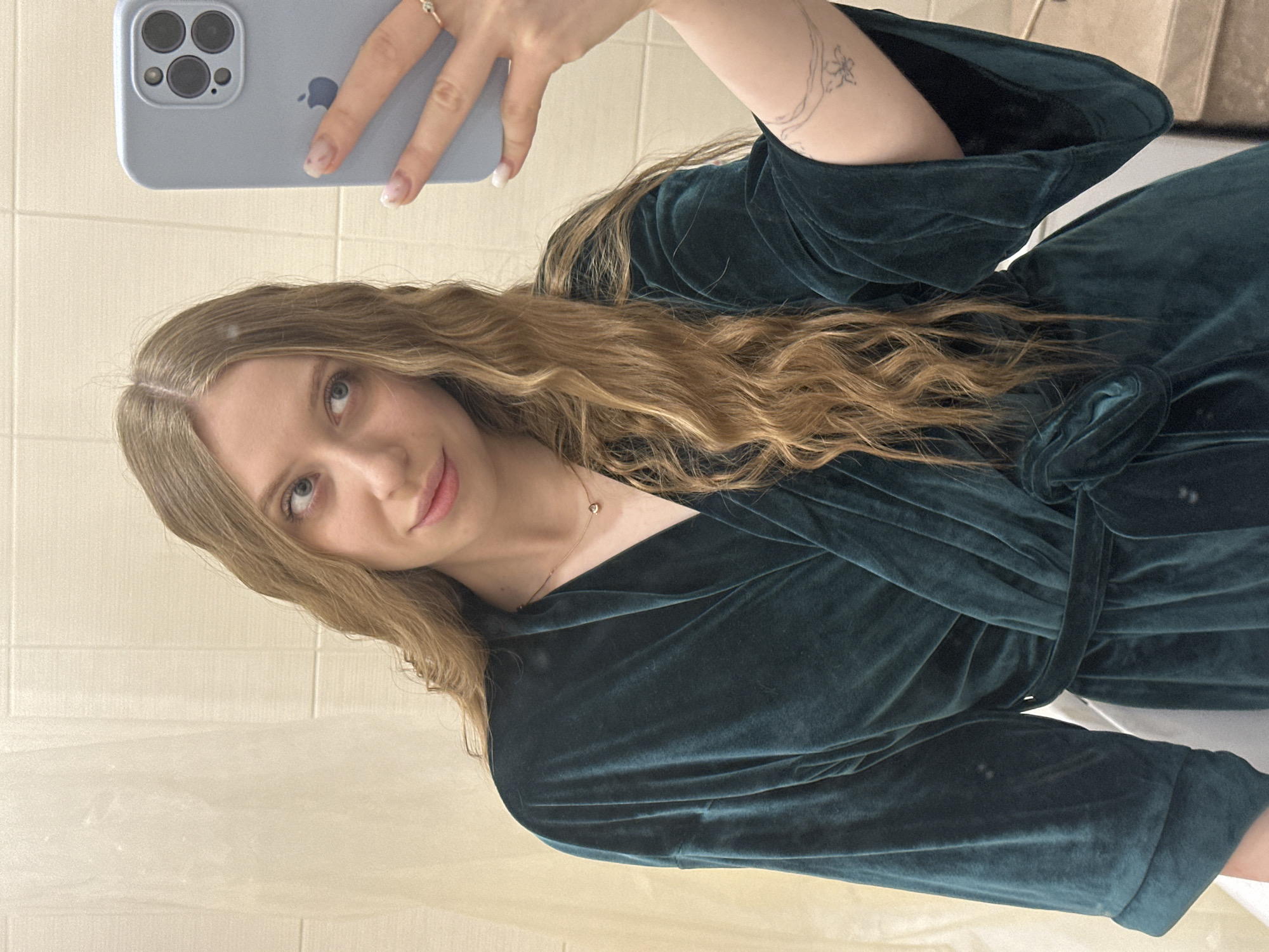 A woman with long, wavy blonde hair and blue eyes is taking a selfie. She's holding a blue iPhone in her left hand, the phone's three camera lenses visible. Her nails are painted with a light pink color and white tips. She is wearing a dark green velvet robe with a belt tied around the waist. A small flower tattoo is visible on her left arm. She is wearing a delicate gold necklace. The background appears to be a bathroom with a light-colored wall and a white curtain or shower liner partially visible. There is no information provided that can locate the location of the subject in the city of Chaikovsky, Russia.