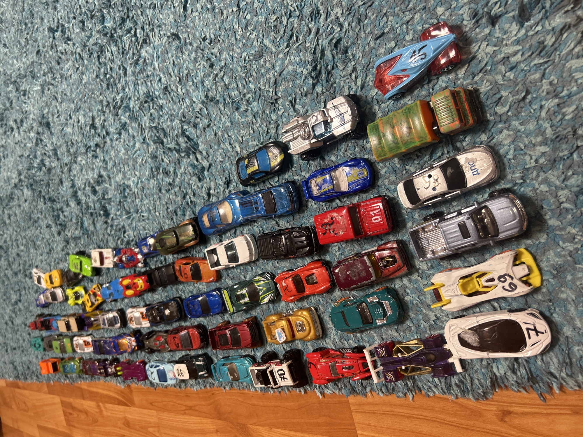 The image shows a collection of approximately 50 toy cars arranged on a blue shag carpet. The cars are of various colors and designs, suggesting a diverse collection of Hot Wheels or similar die-cast models. The cars are arranged in a rough, ascending diagonal line from the bottom left to the top right of the frame. Below the carpet is a section of light-colored hardwood flooring. The scene is indoors. There is no evidence to suggest the location is Izhevsk, Russia. There are no people present.