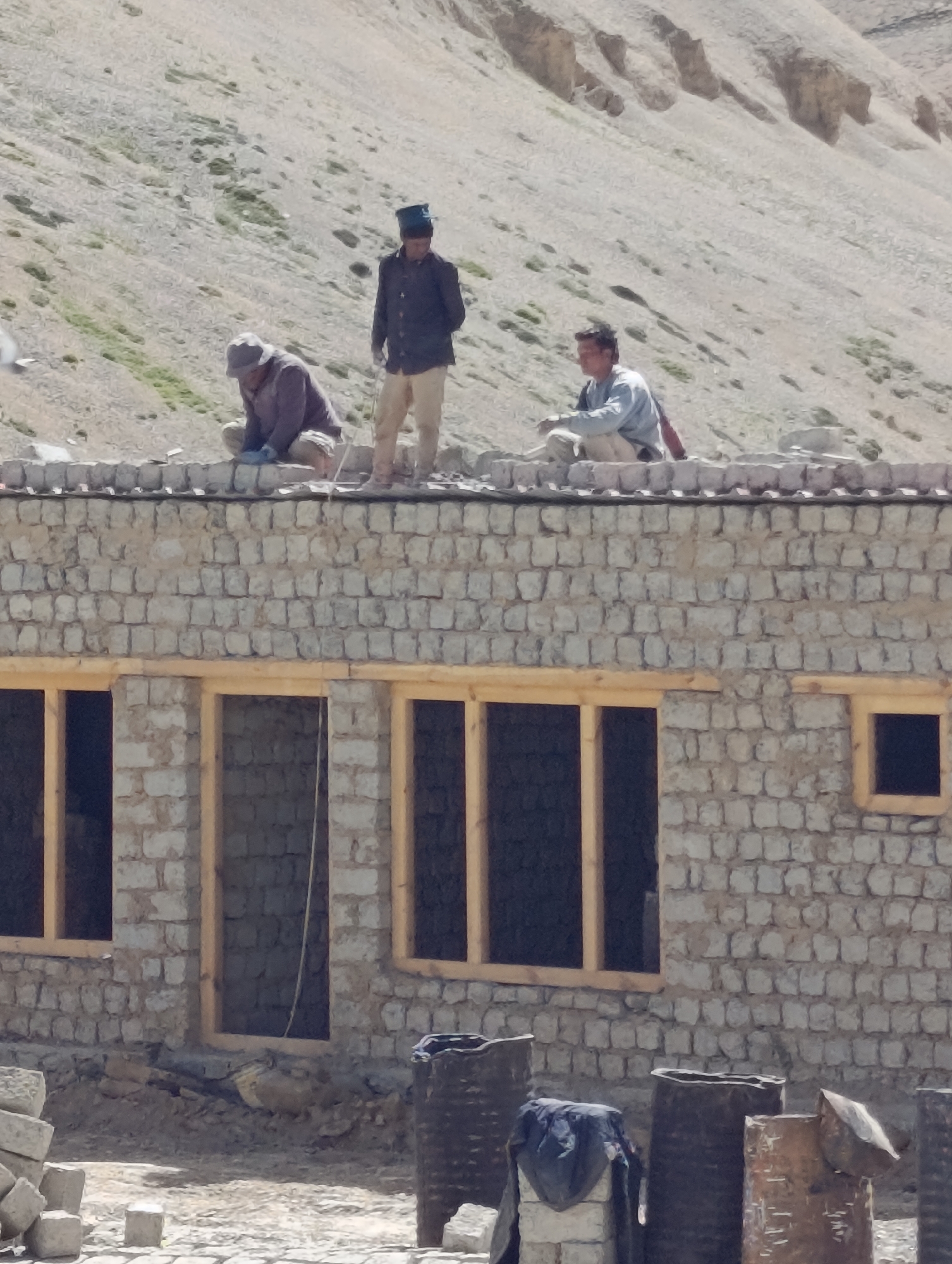 Three men are on the roof of a partially constructed building in Pang, India. The building is made of stone blocks, with wooden frames for windows. The man in the center is standing and wearing a dark jacket and a hat. To his left, another man is crouched down, working on the roof. To his right, a third man is sitting. The background is a steep, arid hillside. In the foreground, there are several large cylindrical objects and stacks of stones.