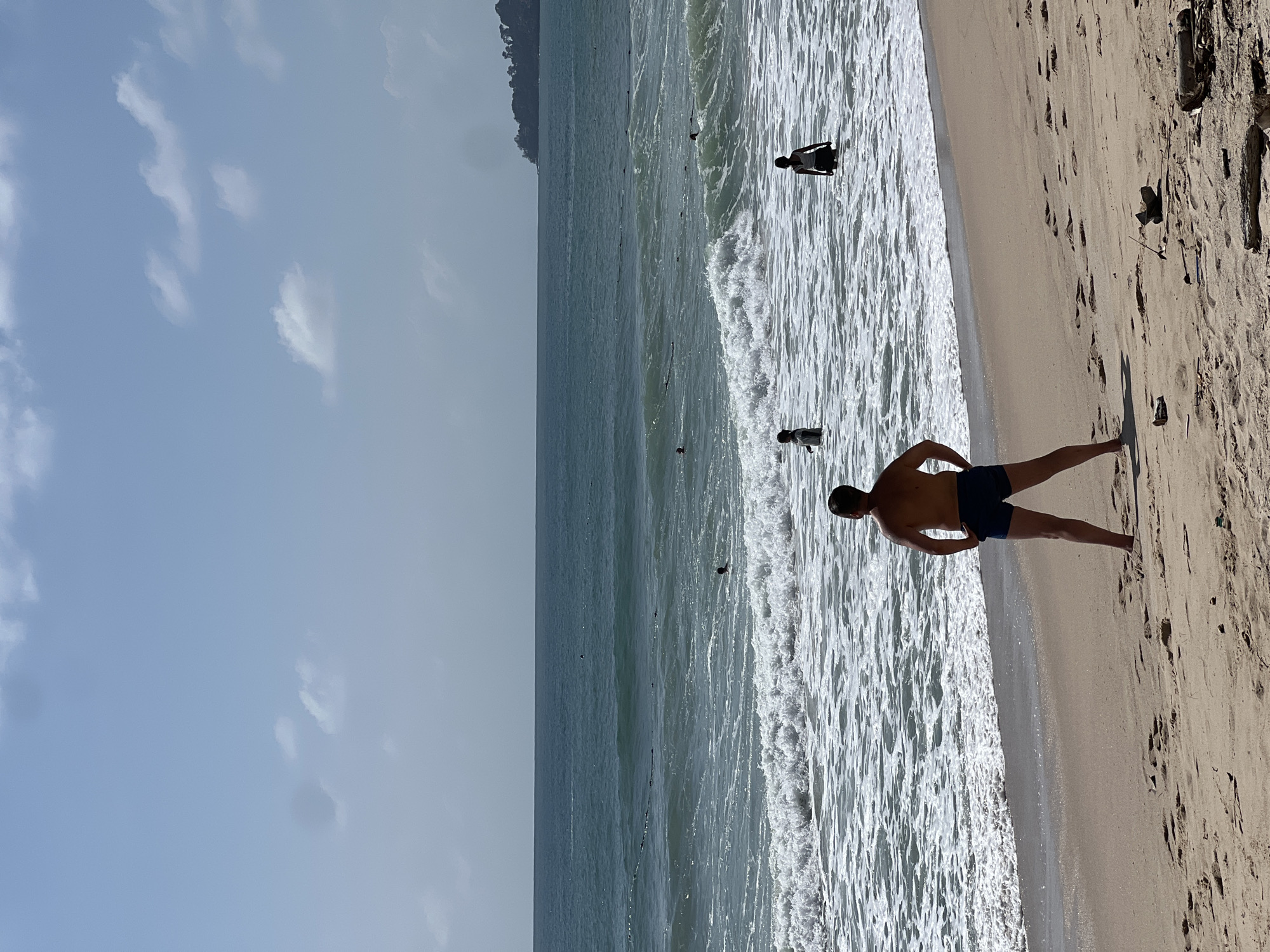 A man stands on a sandy beach in Choeng Thale, Thailand, facing the ocean. He is wearing dark blue swim trunks. In the background, several people are in the water, some swimming and others wading. The sea is turquoise with white waves crashing on the shore. The sky is pale blue with scattered clouds. The beach has footprints and some driftwood. A tree-covered headland is visible in the distance on the right.