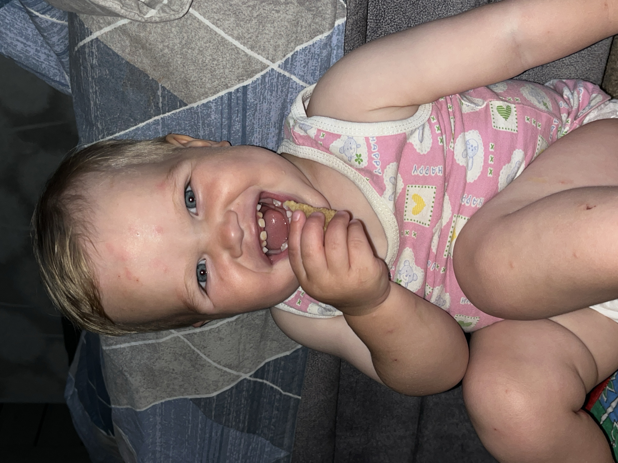A baby lies on a bed with a blue and gray patterned sheet. The baby is wearing a pink and white onesie with cartoon animal designs. The baby is fair-skinned with blonde hair and blue eyes, and has a snack in their hand. The baby is smiling, showing their teeth. Red spots are visible on the baby's face. The location cannot be determined from the given image.