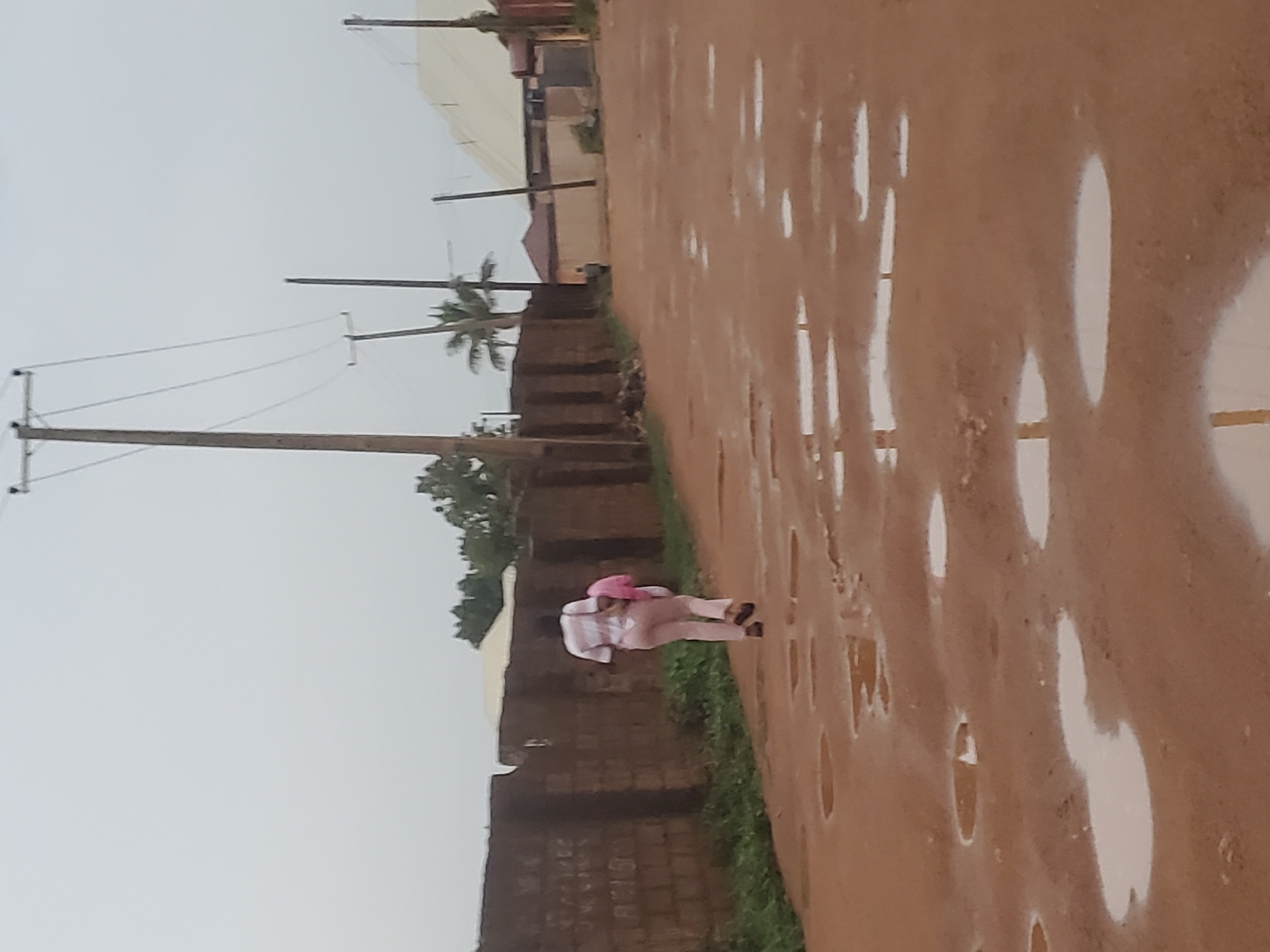 A medium shot of a street in Jos, Nigeria, shows a clay road filled with puddles. A person wearing light-colored pants and a white shirt is walking down the road. The person is wearing a pink hat. A brick wall lines the left side of the road. Power lines run above the wall and road. The sky is overcast.