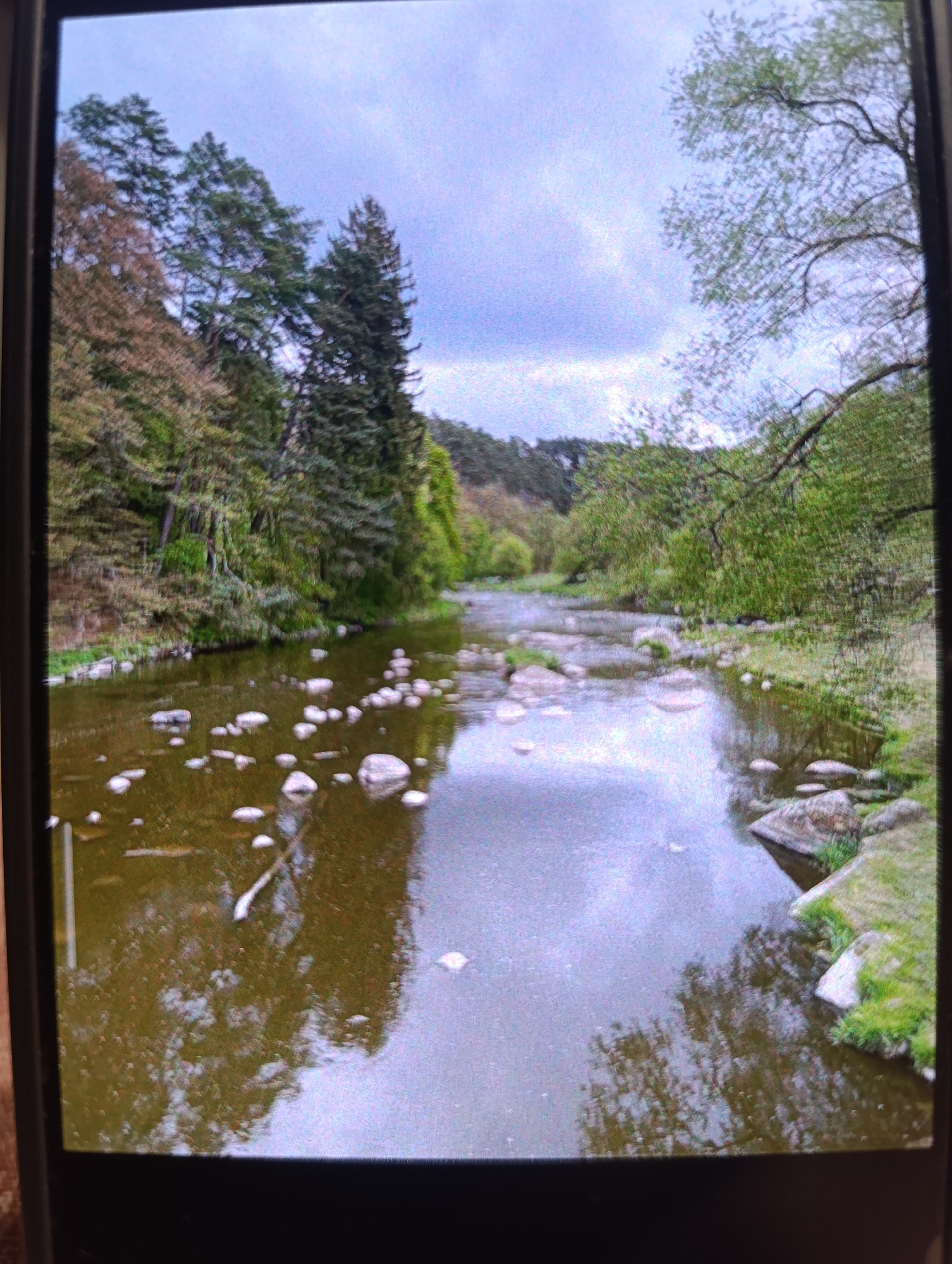 The image shows a river with rocks scattered in its shallow water. The riverbanks are lined with lush green trees and foliage. The sky is overcast with clouds, creating a subdued light. The overall scene is serene and natural, suggesting a peaceful outdoor environment. The image is taken from a slightly elevated angle, providing a good view of the river and its surroundings. The water reflects the sky and trees, adding depth to the scene. There are no people or animals visible in the image. The location is an unknown city in an unknown country.