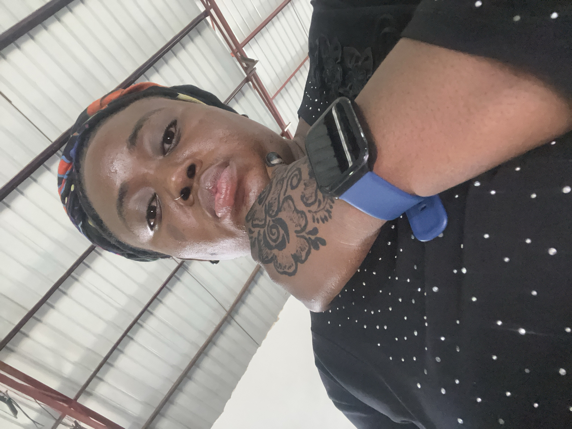 A selfie of a dark-skinned person with a headscarf is oriented so the camera faces down. The individual has a tattoo on their neck and wears a smartwatch with a blue band. They are wearing a black garment with small white dots. The background is a ceiling with exposed beams and white paneling. The location is Barakin Akawo, Nigeria.