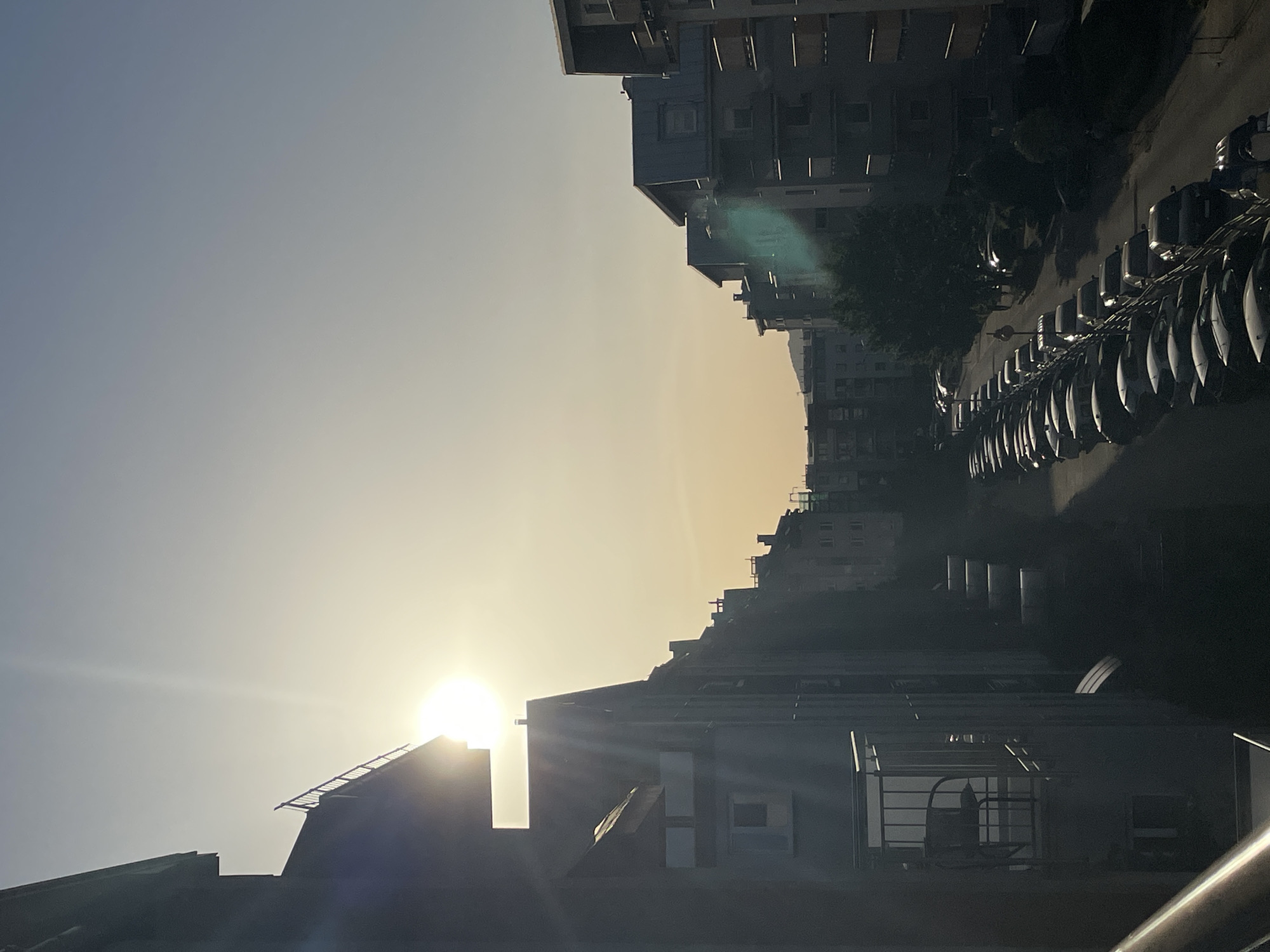 The image shows a street scene in an urban environment at sunset. Several apartment buildings line the street. A row of parked cars is visible on the right side of the street. The sun is setting behind a building in the foreground, casting a glare and creating a silhouette effect on some of the buildings. The sky is a gradient of pale blue to orange as it approaches the horizon. The exact location is unknown.