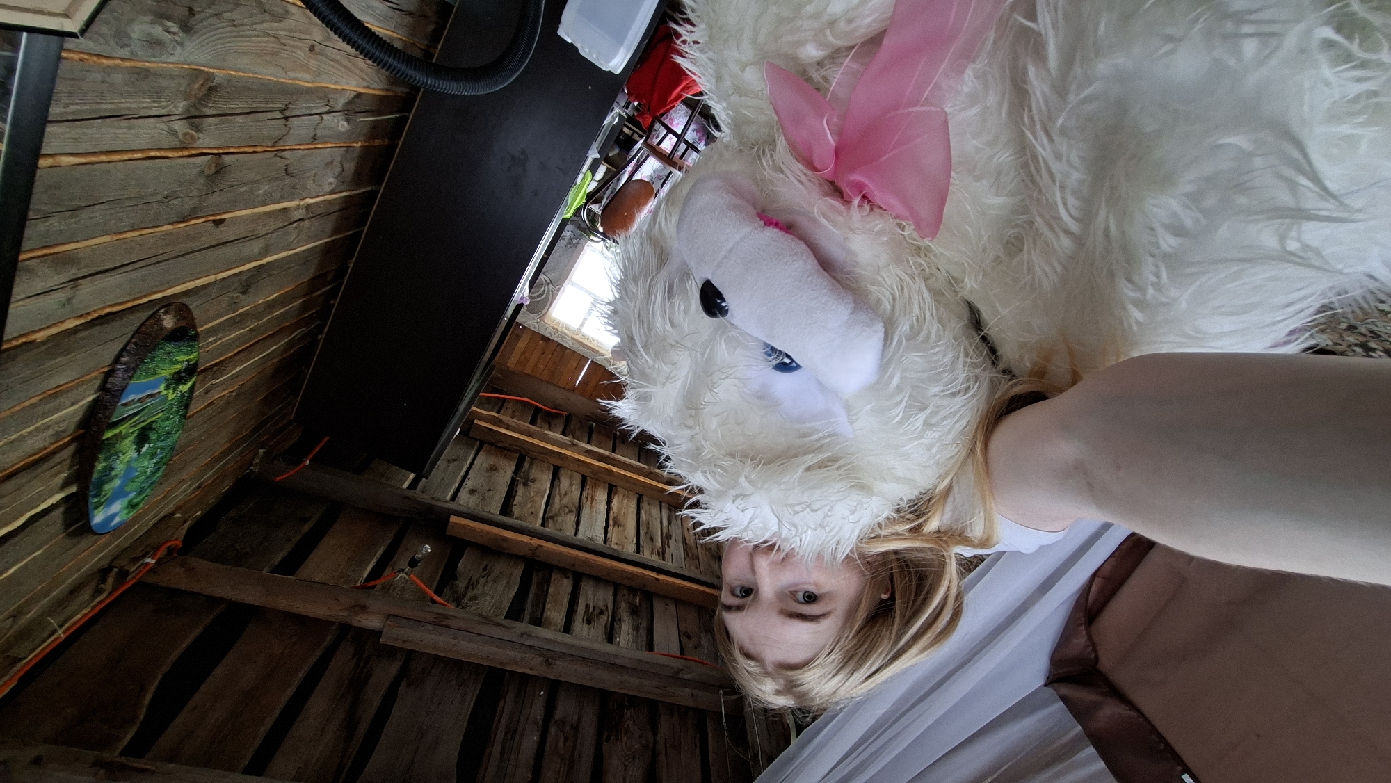 The image presents a disorienting, upward-angled perspective from within a wooden structure in Большой Букор, Russia. A woman, partially visible, is positioned below a large, white, fluffy stuffed animal with a pink bow. The stuffed animal dominates the upper half of the frame. The woman's face is visible, suggesting she is holding the stuffed animal above her.
The environment consists of a rustic wooden interior, characterized by roughly hewn planks forming the walls and ceiling. An oval picture is hung on the wooden wall with a picture of greenery on it. Some electrical wires are visible. A dark-colored, possibly metal, structure or shelf is positioned at the top-left of the frame. A partial view of other objects are visible in the top of the frame near a window. The lighting appears to be a mix of natural light from a window and artificial light within the structure. A draped fabric is visible below the woman.