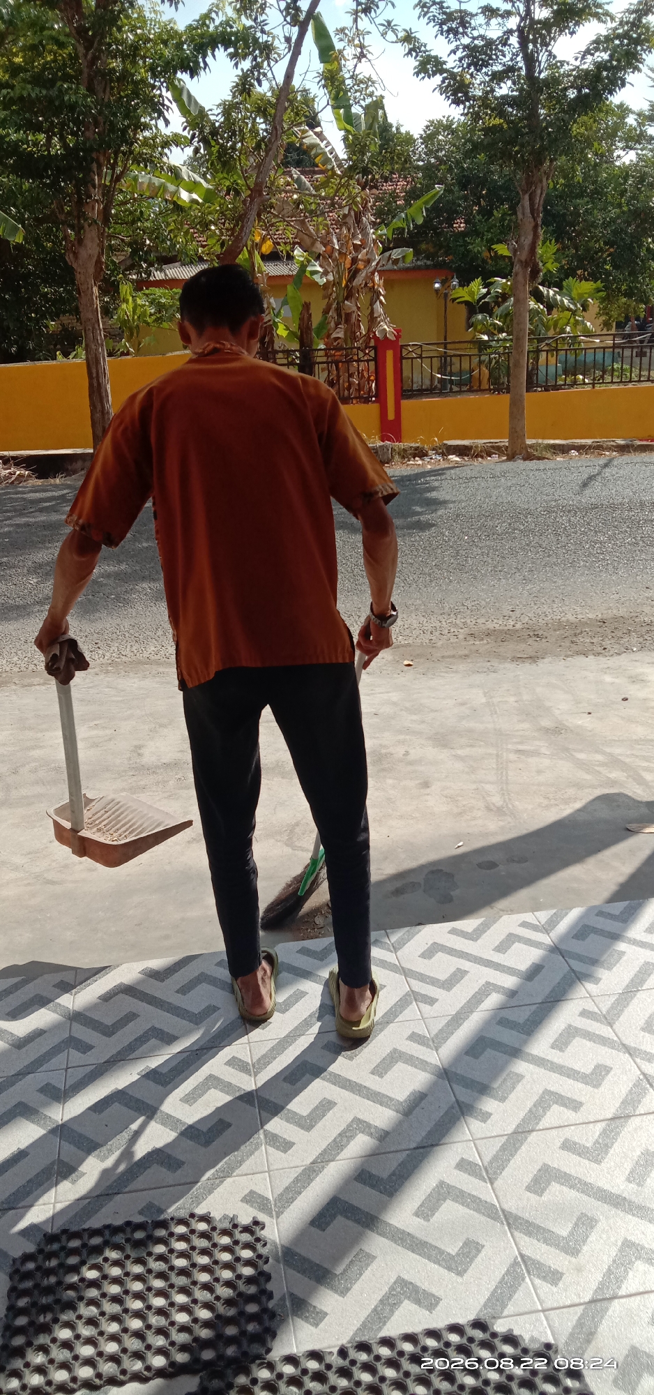 In Pakong, Indonesia, on August 22, 2026, at 08:24, a person is standing on a patterned tiled surface, holding a dustpan in their left hand and a broom in their right hand. The person is facing away from the camera, wearing a short-sleeved orange shirt and black pants. The background shows a yellow wall, a black fence, and trees. The ground is a mixture of pavement and tiles, with shadows cast by the sun.