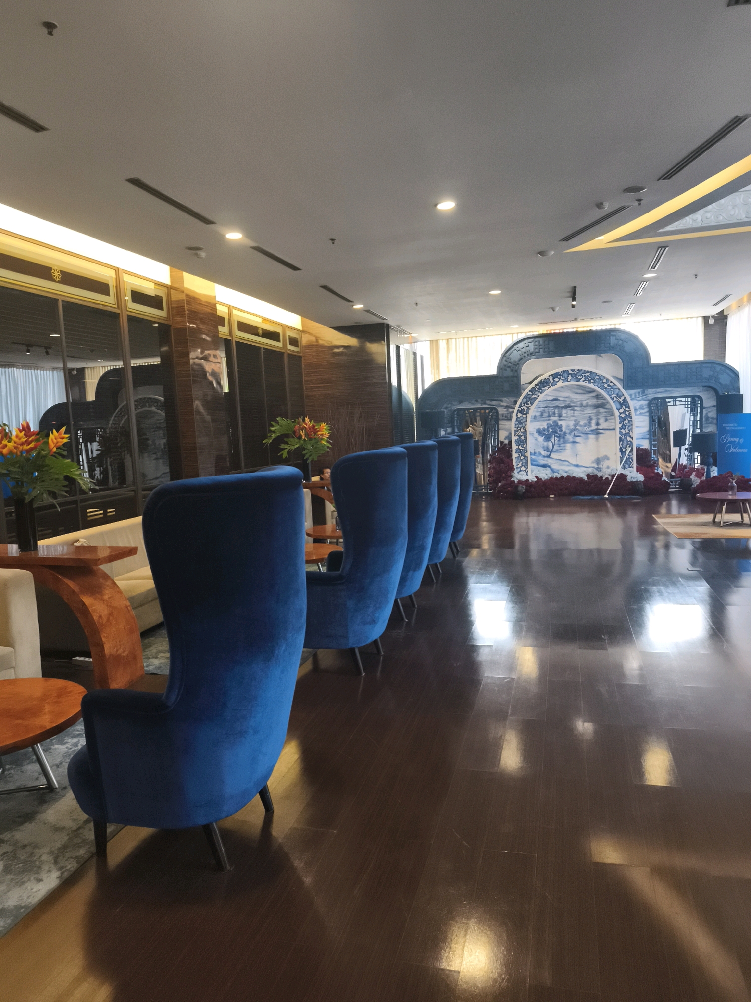 The image depicts a well-lit indoor event space, possibly a banquet hall or a reception area, situated in Palembang, Indonesia. The environment is characterized by a polished wooden floor that reflects the overhead lighting. In the foreground, a row of plush, royal blue velvet armchairs is arranged facing towards a decorative archway in the background. This archway is embellished with a blue and white landscape mural, possibly depicting a traditional Chinese scene, and framed by dark red floral arrangements.
To the left of the armchairs, there are modern side tables with orange tabletops and what appears to be a light-colored sofa in the background. Two vases filled with vibrant orange and yellow tropical flowers add a splash of color to the scene. Further back, to the right of the main archway, a sign reading "Welcome to the Beginning of Togetherness" is visible, suggesting a wedding or a similar celebratory event. The ceiling features recessed lighting and ventilation systems, indicating a modern interior design. The overall ambiance is elegant and festive, designed to host a special occasion.