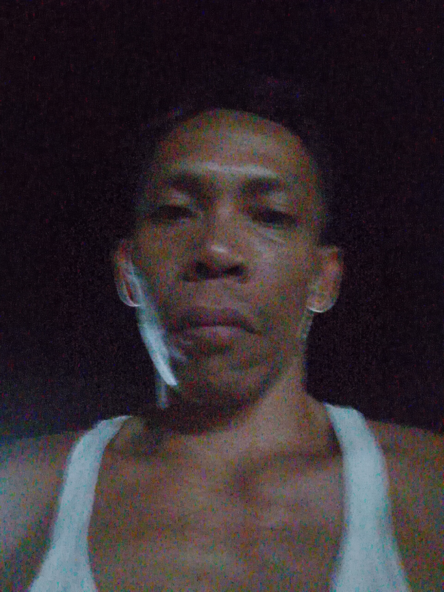 A man with short hair and dark skin is facing the camera. He wears a white tank top and earrings. The background is dark. It is impossible to determine the location is Bukit Ketok, Indonesia, without further information.