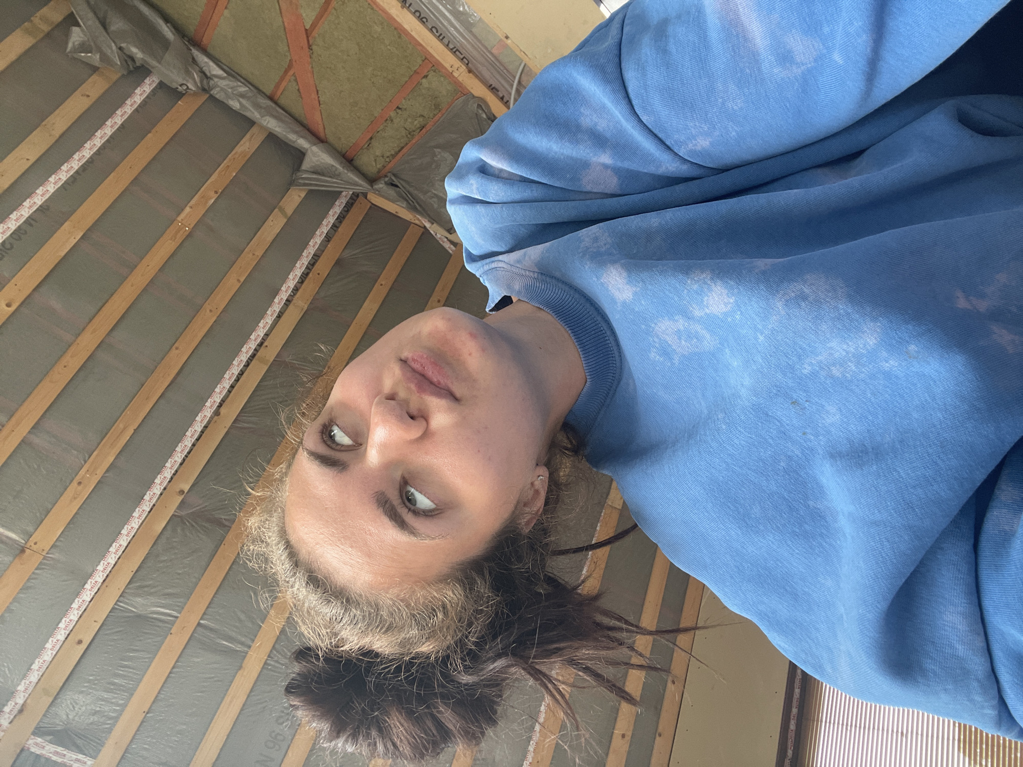 The image shows a close-up, low-angle shot of a young woman looking upwards and to the right. She has her hair pulled up in a messy bun and is wearing a blue tie-dye sweatshirt. The background appears to be the underside of a roof under construction, with wooden beams and insulation visible. There is no indication of the specific location within Мосметаллоконструкция Онт, Russia.
