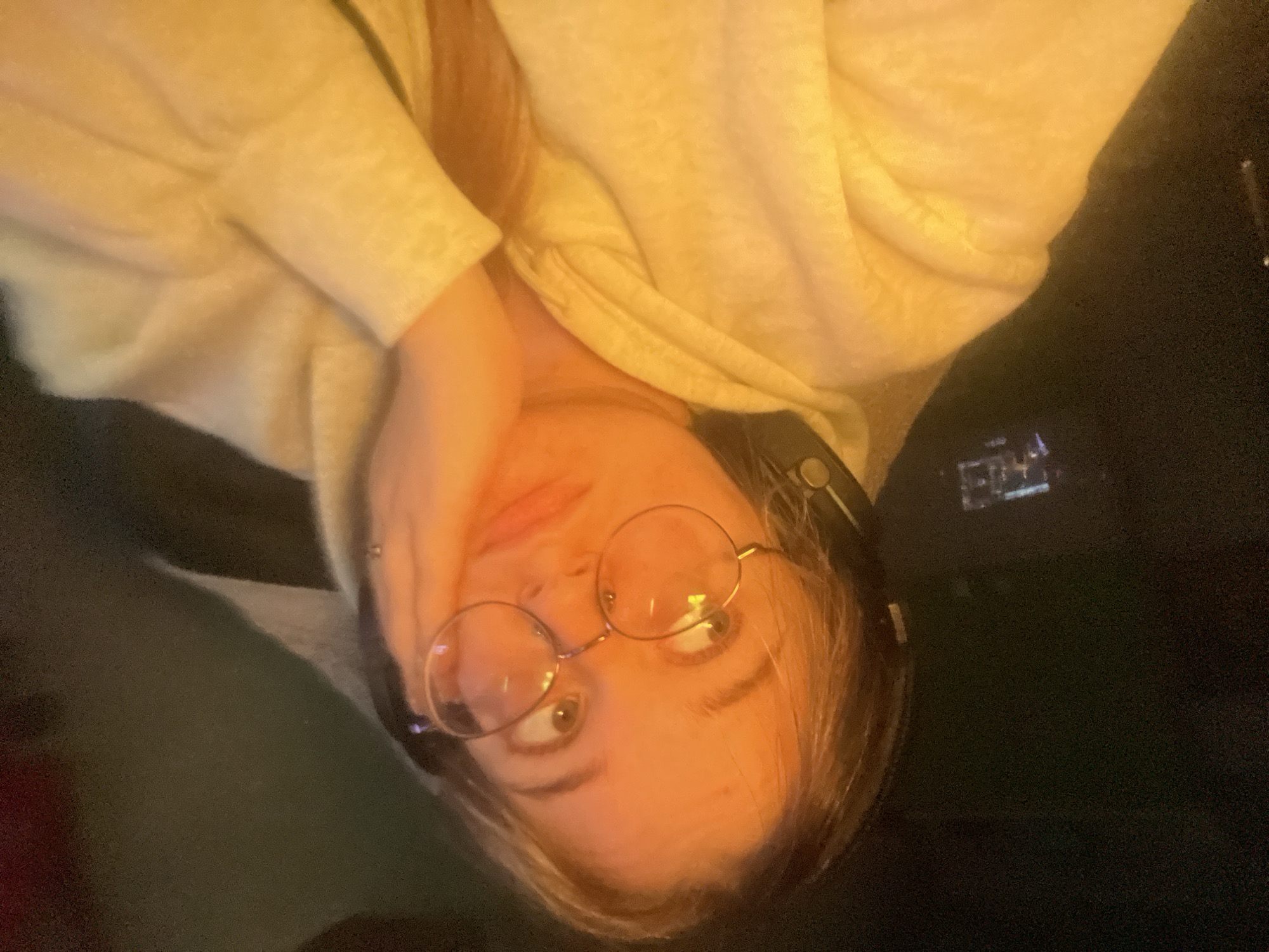 A young person with blond hair and round glasses is looking off to the side with their hand on their cheek. They are wearing a light-colored hooded sweatshirt and headphones. The background is dark, with a small screen visible on the left side. The image appears to be a selfie taken indoors. There is no identifiable information that would suggest a location in Kolomna, Russia.