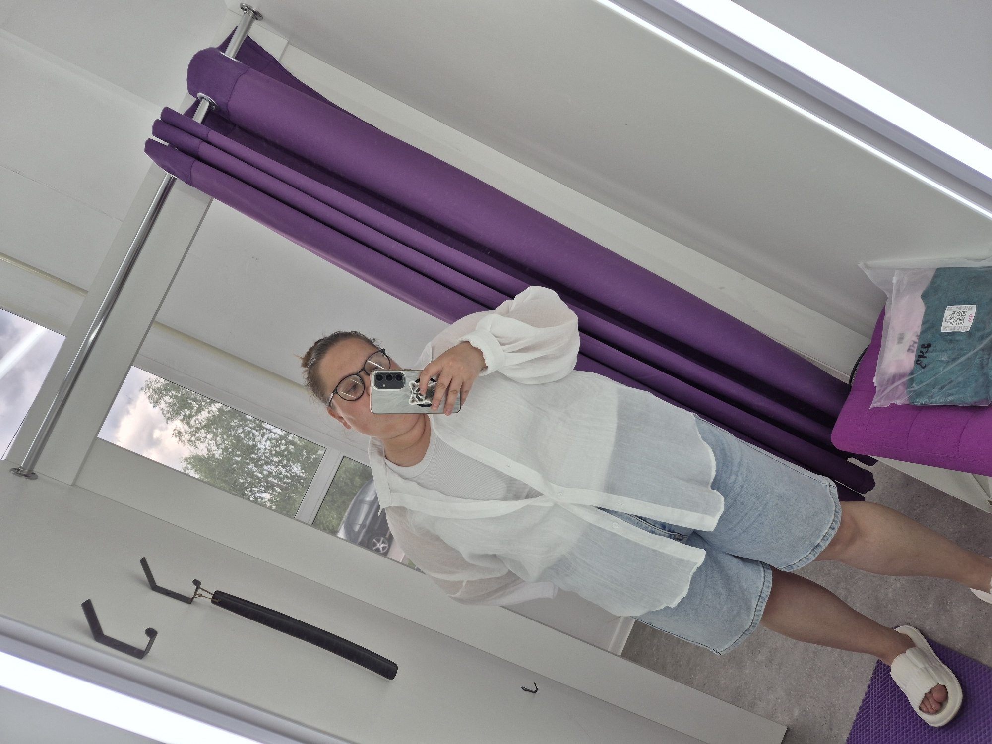 The image is a selfie taken inside a dressing room, likely in a retail store. A person with glasses is holding a phone up to take the picture. They are wearing a white top over a white t-shirt and light blue shorts, and white slide sandals. A purple curtain hangs behind them. There's a clothing rack hook on the wall to the left. A window is visible in the background with trees visible. The package is located on the purple bench in the upper right. The image was not necessarily taken in the city of Chaikovsky, Russia.