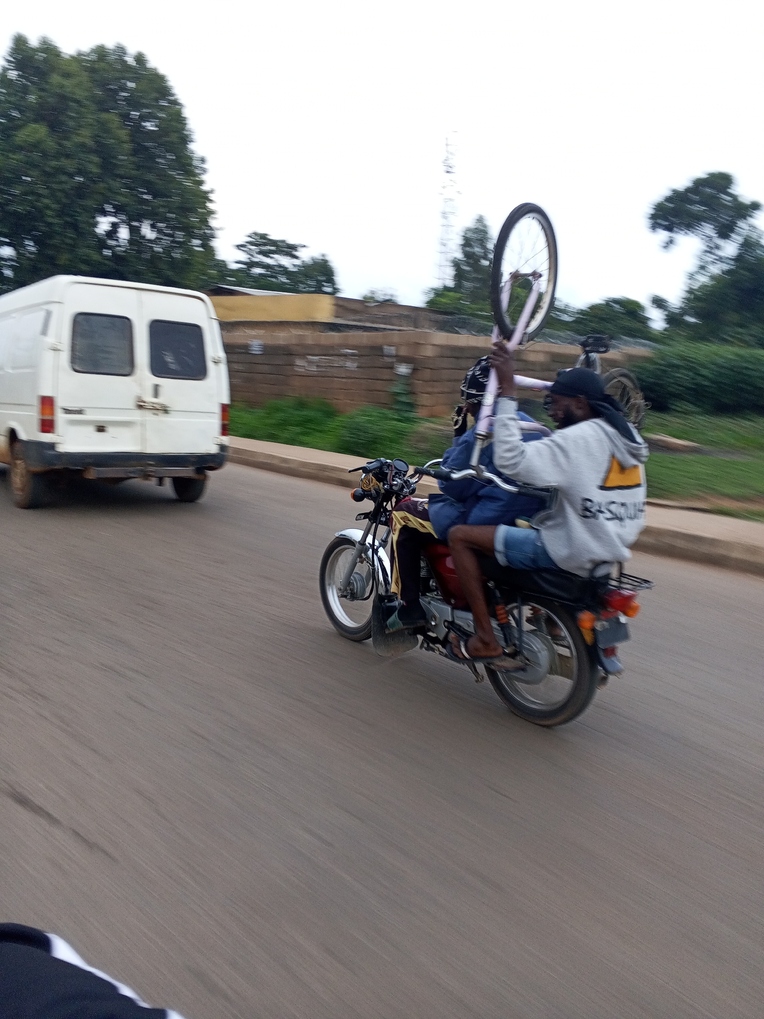 In Jos, Nigeria, two individuals are traveling on a red motorcycle. The person in the rear is holding a bicycle vertically. The motorcycle is moving on an asphalt road, and a white van is visible in the background. The environment appears to be a semi-urban area with trees and buildings. The lighting suggests it is daytime, possibly overcast. The motion blur indicates the vehicles are in motion.