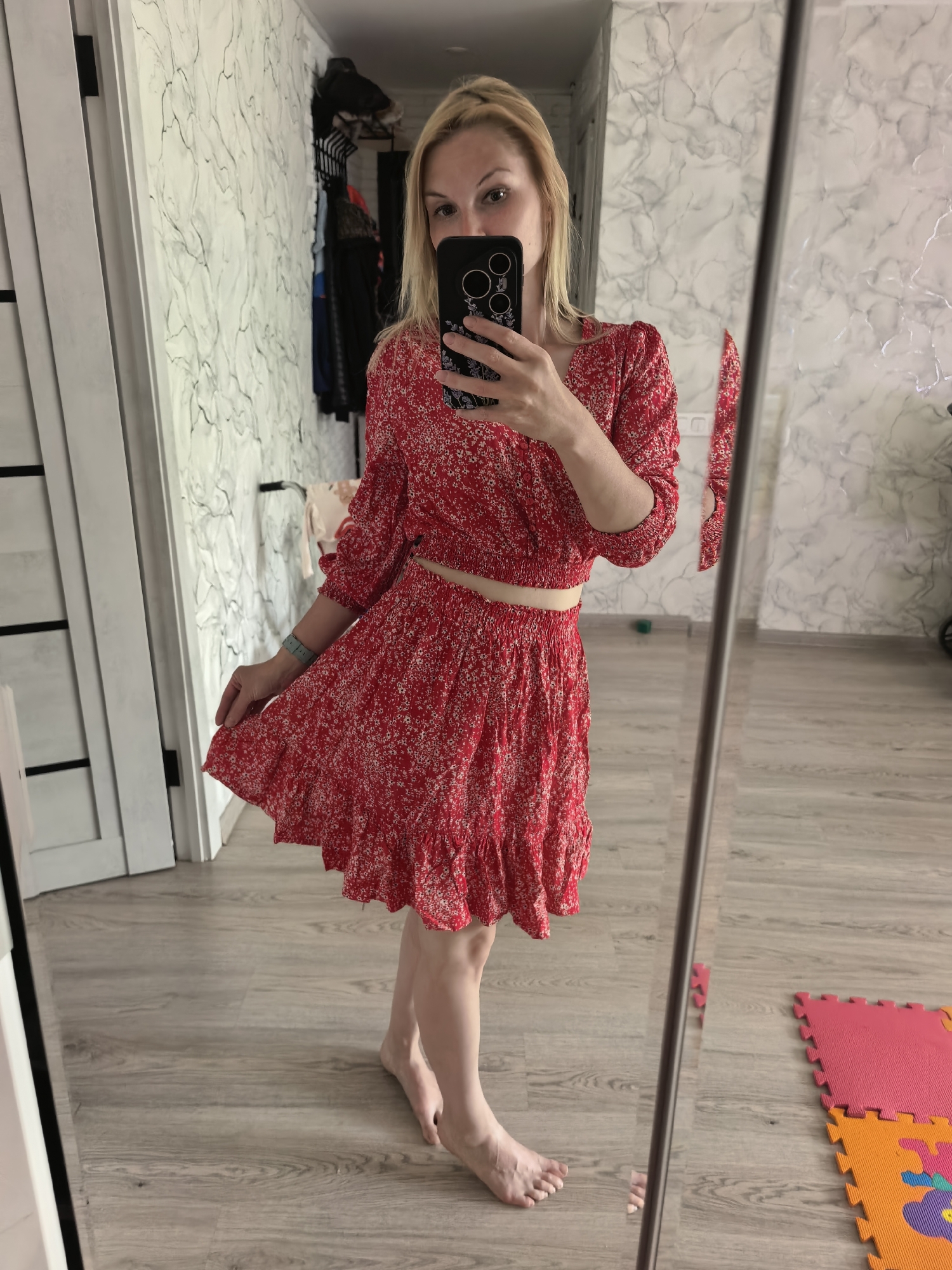 A woman stands barefoot in front of a full-length mirror, holding a smartphone to take a selfie. She is wearing a two-piece red floral dress with long sleeves and a bare midriff. The room is a domestic interior with a light-colored wood floor and white walls with a marble-like pattern. A doorway is visible to the left, and a clothes rack hangs against the wall. A child's foam play mat is in the lower right corner. The photo appears to be taken indoors. It is not possible to verify that the location is in the city of Sterlitamak, Russia.