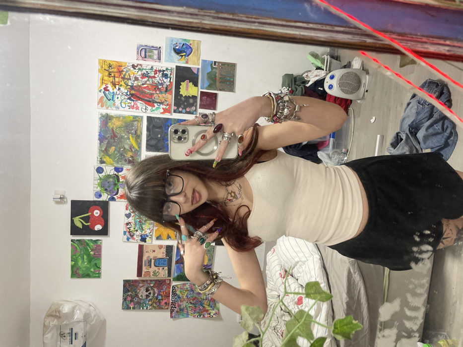 A person with brown bangs, wearing black-framed glasses, a white tank top, and a black skirt, is taking a selfie in front of a mirror. Their hands are adorned with numerous rings and bracelets. The background features a white wall decorated with colorful drawings and paintings. A bed with patterned sheets is visible in the lower portion of the frame, and a clutter of items, including a small white heater, is seen in the upper right corner. A plant with green leaves is partially visible in the foreground.