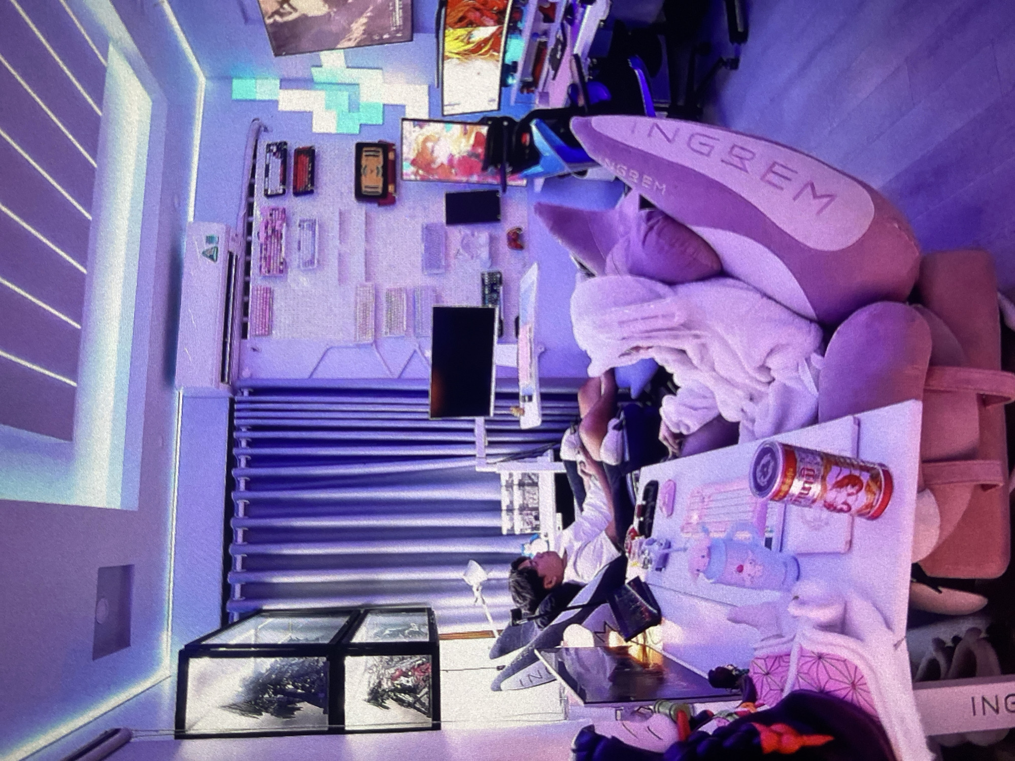 A room appears to be located in Lhokseumawe, Indonesia. It includes a person sitting at a desk cluttered with various objects, including a drink can with an anime character, stationery, and small figures. The seating arrangement seems to accommodate a gaming or computer setup, with multiple monitors visible on the wall along with decorative items and keyboards. The room's lighting is dominated by a blue hue, possibly from LED lighting. Additional details in the room include what seem to be storage containers, some featuring artwork or designs. A large, plush headrest is positioned on the chair, suggesting a focus on comfort or extended use.