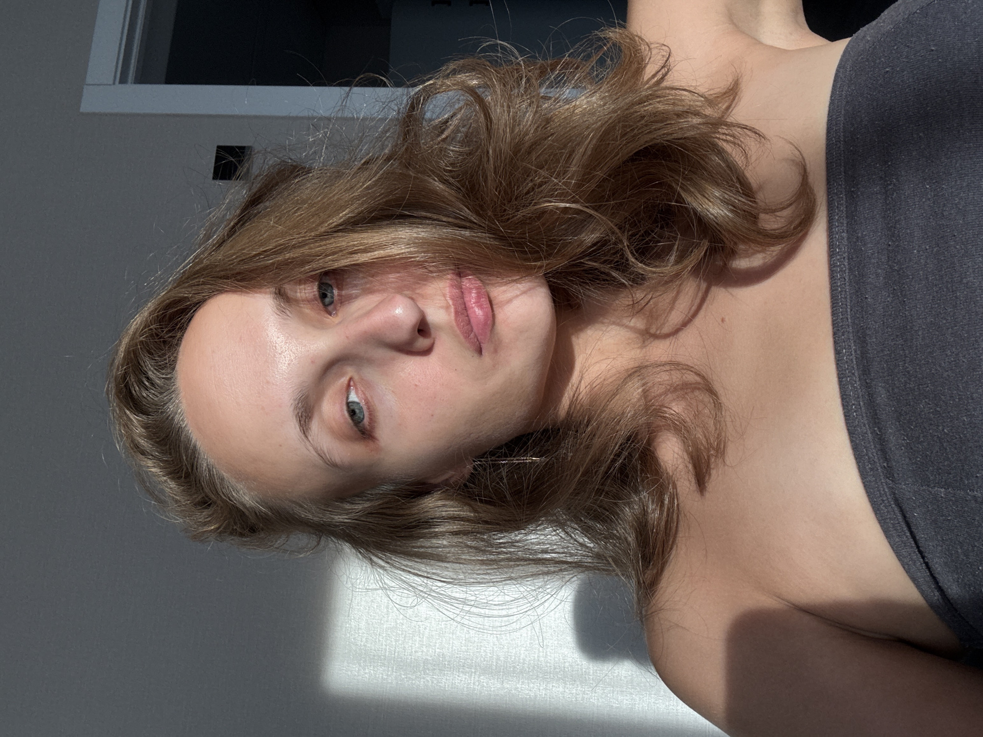A young woman with light brown hair is photographed from the chest up, looking directly at the camera with a neutral expression. She is wearing a dark gray strapless top. The lighting suggests a bright, sunny day with strong shadows cast on the wall behind her, indicating she is indoors near a window. The background appears to be a plain wall with a doorway visible on the right side. There is no information provided that would allow for the determination of the location within the city of Подшивалово, Russia.