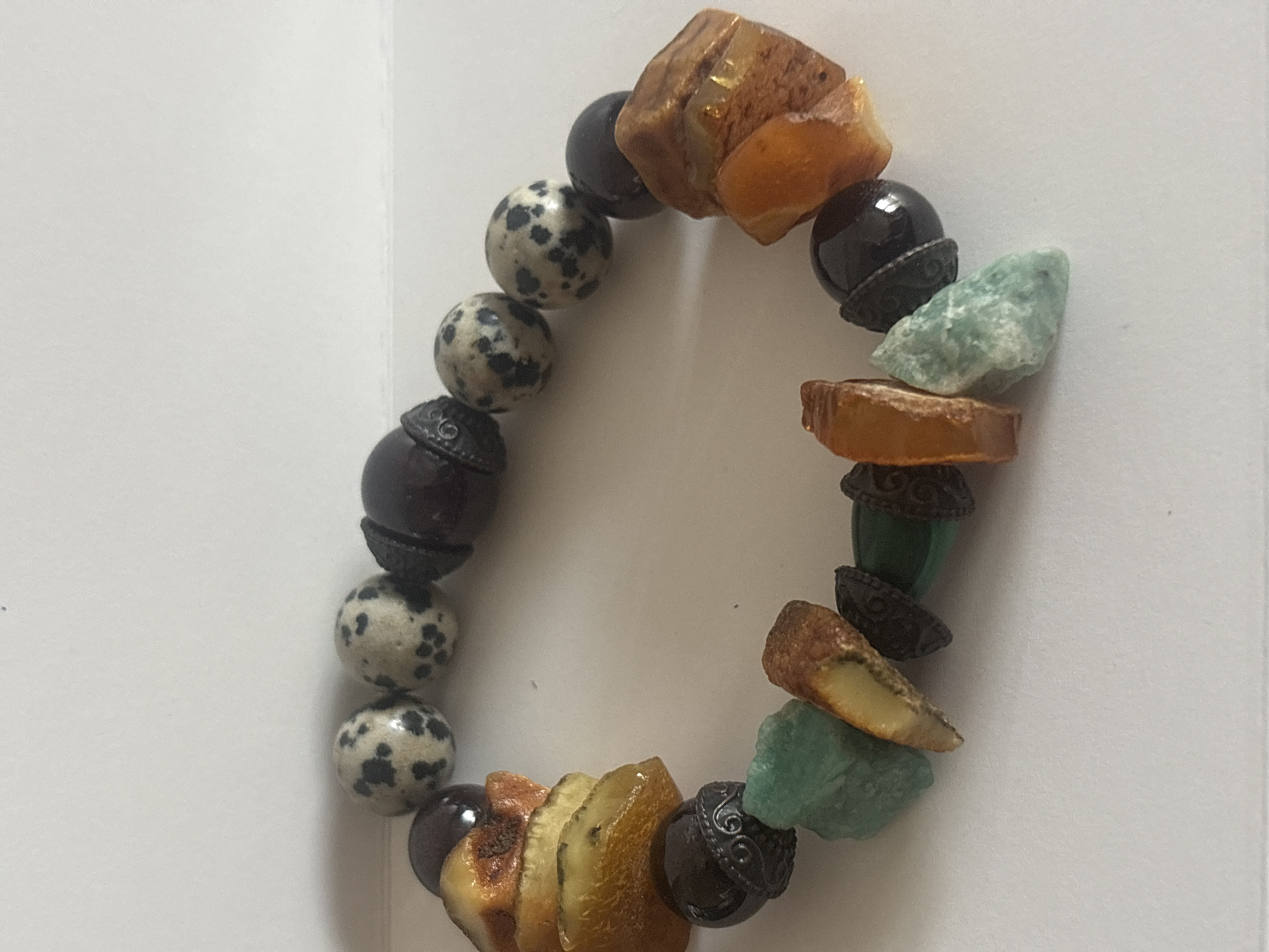 An overhead, close-up shot captures a colorful bracelet laid in an oval shape against a plain white background. The bracelet features a diverse array of beads and natural stones. Prominent elements include polished spherical black beads and spherical white beads with black speckles, resembling dalmatian jasper. These are interspersed with irregularly shaped, raw pieces of translucent amber, ranging in color from golden yellow to warm orange-brown. Rough-cut, light green-blue stones, possibly aventurine or amazonite, and some cylindrical dark green beads also form part of the design. Dark metallic spacer beads, adorned with intricate embossed patterns, separate many of the stones, adding an ornate detail. The lighting is bright and even, highlighting the natural variations and textures of each bead. No activity is depicted, and there is no visible text or emotional context. This item is located in Bar, Montenegro.