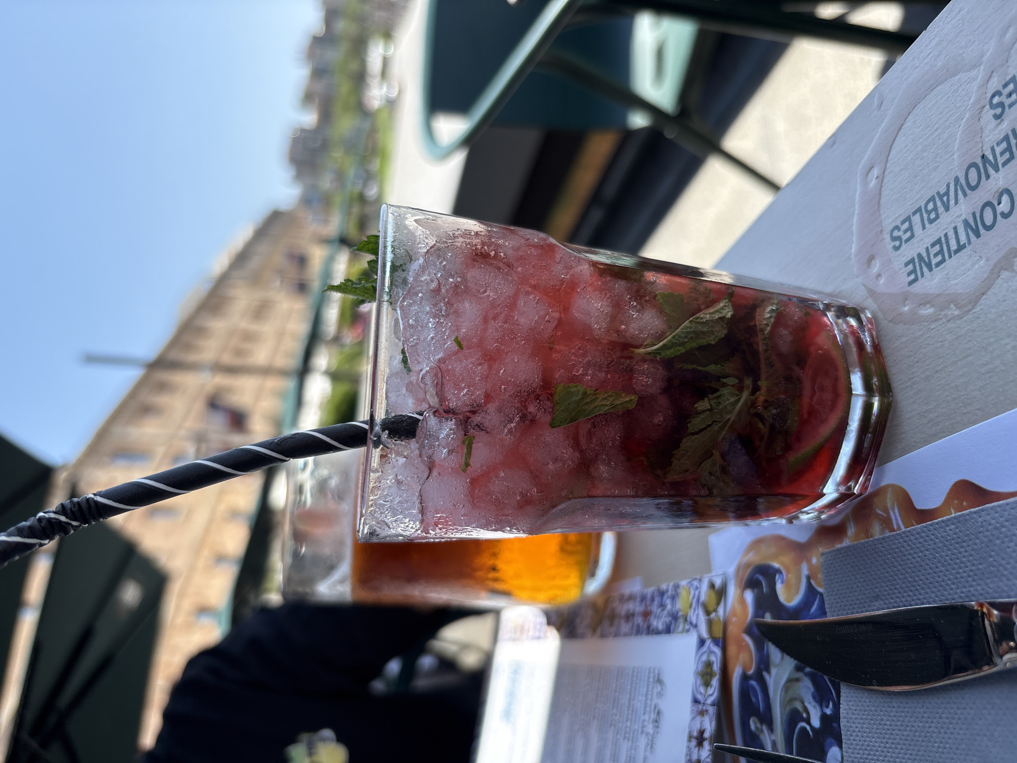 The image shows a refreshing cocktail on a table in Barcelona, Spain. The drink is in a glass, filled with ice, a red liquid (possibly juice or a mixed drink), and mint leaves. There's a black and white striped straw in the drink. The cocktail is sitting on a table that has a menu or placemat with a blue and white pattern. A napkin and silverware are also visible near the drink. In the background, out of focus, is a building.