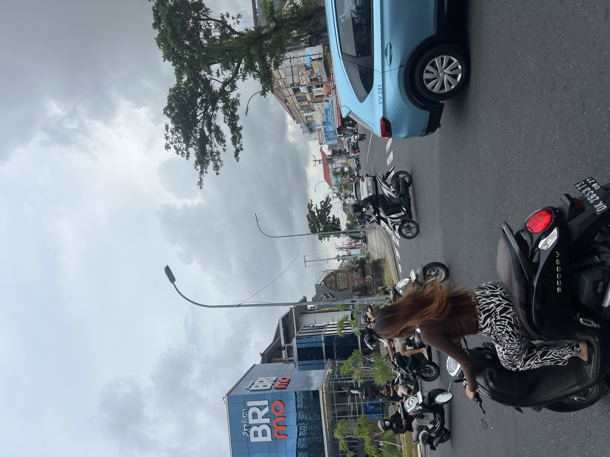 The image shows a street scene in Kuta, Indonesia, under an overcast sky. In the foreground, a woman with long brown hair is seated on a black scooter, wearing a brown top and zebra-print pants. Several other scooters are parked behind her, some with riders wearing helmets. A blue car with the inscription "VV 231" is positioned at the top of the frame, partially obscuring the view of the road ahead. A BRImo bank building is visible on the left side of the image. The street is lined with buildings, trees, and streetlights. Other vehicles and pedestrians are seen further down the road. A scooter registration plate reads "DK 2853 TS 08.27".
