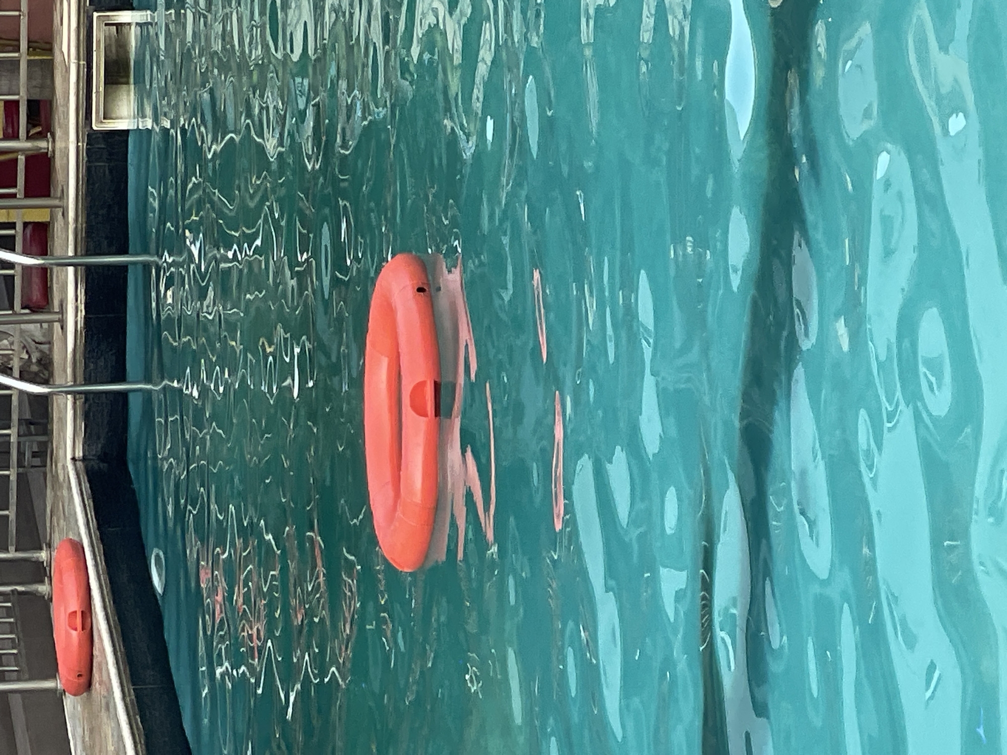 The image shows a swimming pool with clear, teal-colored water. The surface of the water is rippling, creating distorted reflections of the surroundings. Two bright orange life preservers are visible. One is floating in the pool, partially submerged, while the other is resting on the pool's edge, near a metal ladder. The ladder, made of shiny metal, is partially visible on the left side of the image. The scene appears to be outdoors, possibly on a sunny day, given the bright colors and the quality of light reflecting off the water. The overall impression is one of a recreational setting. There is no visible text in the image.